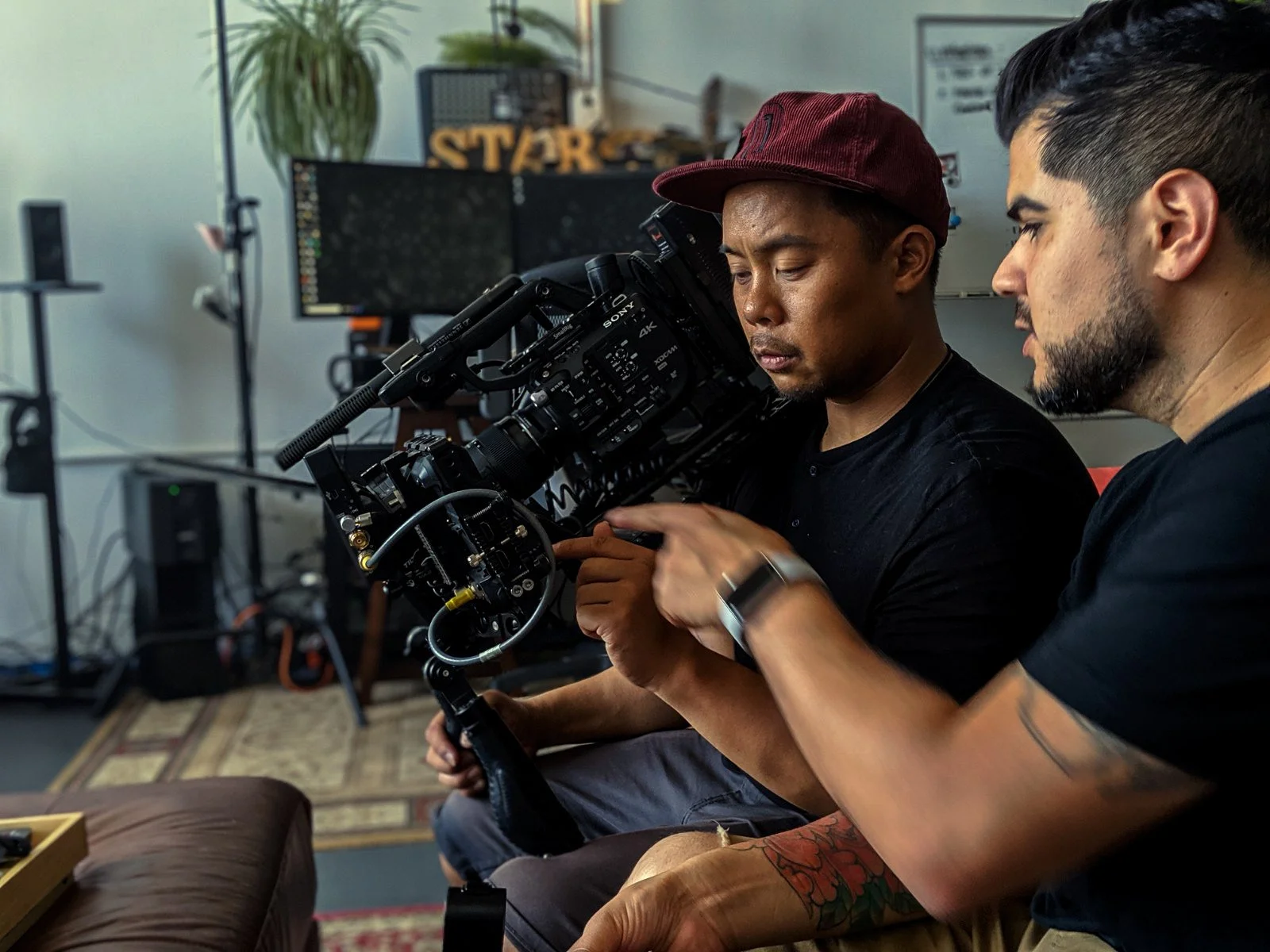 Two men sitting indoors, one holding a professional camera on a stabilizer, reviewing footage while the other points at the camera, both focused on the device.