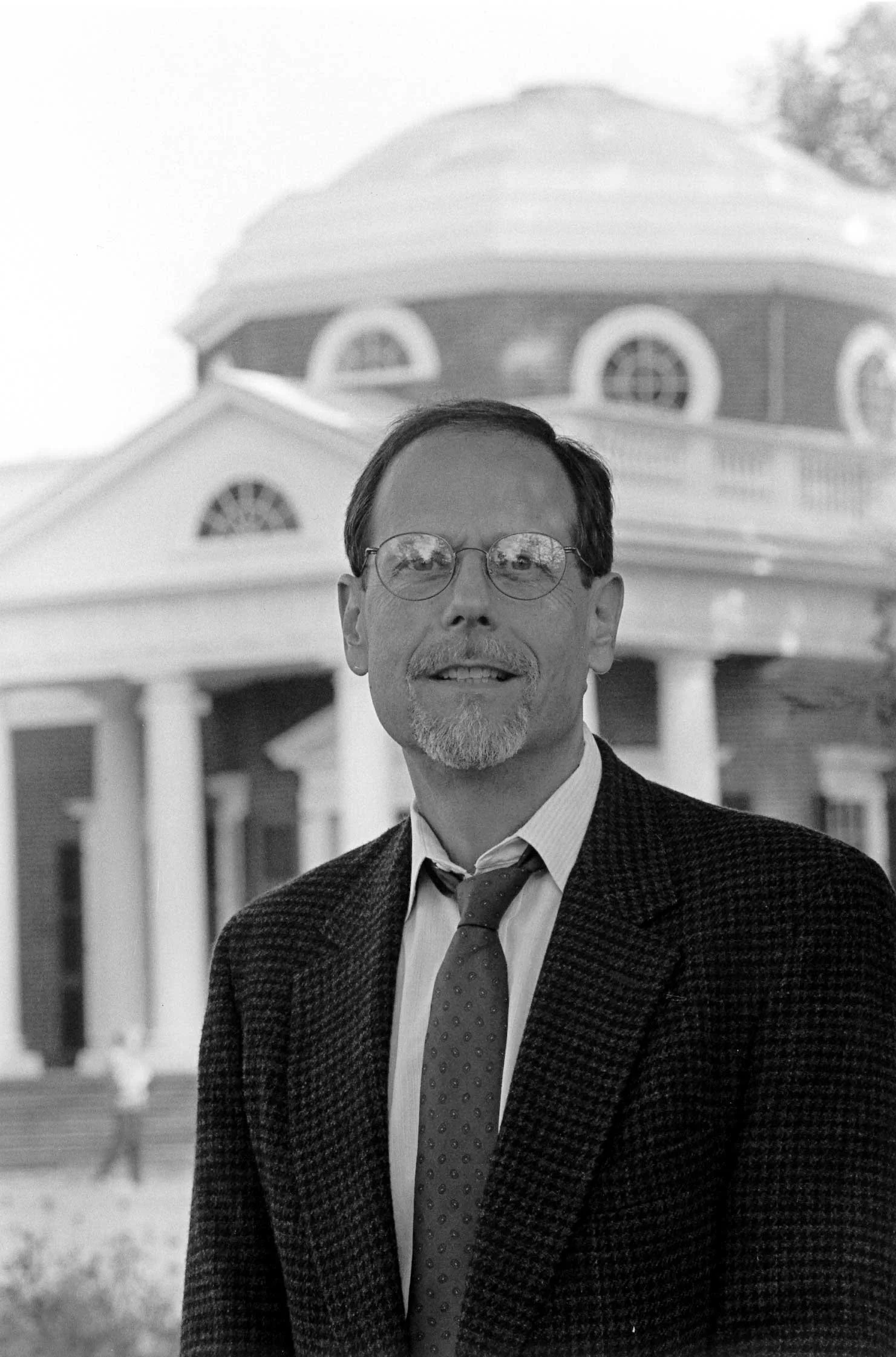 A man with glasses, a beard, and a mustache wearing a suit and tie, standing outdoors in front of a large official-looking building with columns and a dome.