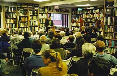 A person giving a lecture or presentation in a small library or bookstore, with an audience seated facing the speaker, surrounded by tall bookshelves filled with books.