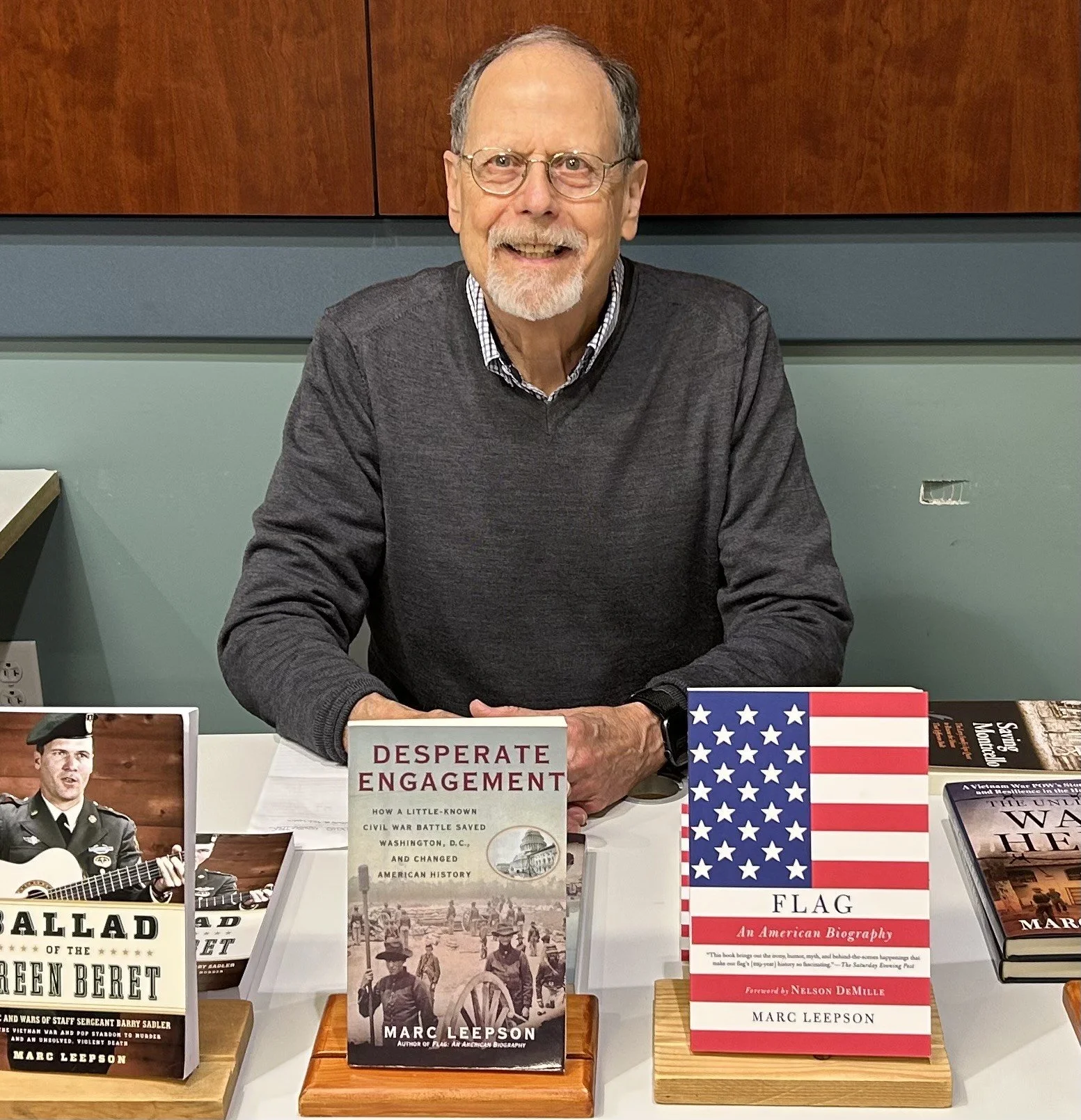 A man with glasses, gray hair, and a beard, smiling, sitting at a table with books and a photo of a soldier playing guitar in front of him.