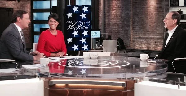 Three TV hosts sitting at a news table on a talk show set with a large screen behind them displaying the words "What So Proudly" and a graphic of the American flag.