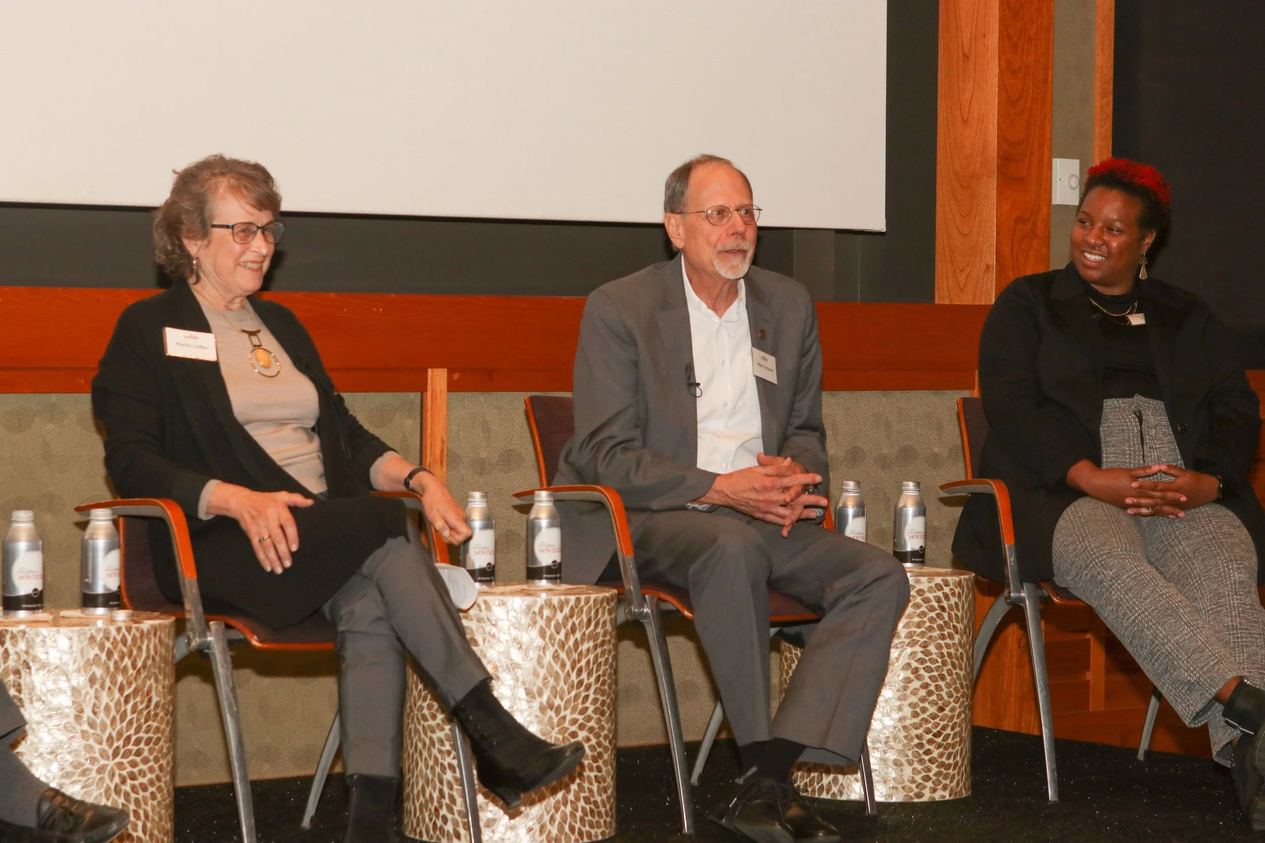 Three people sitting on a panel during a discussion, with water bottles on tables in front of them.