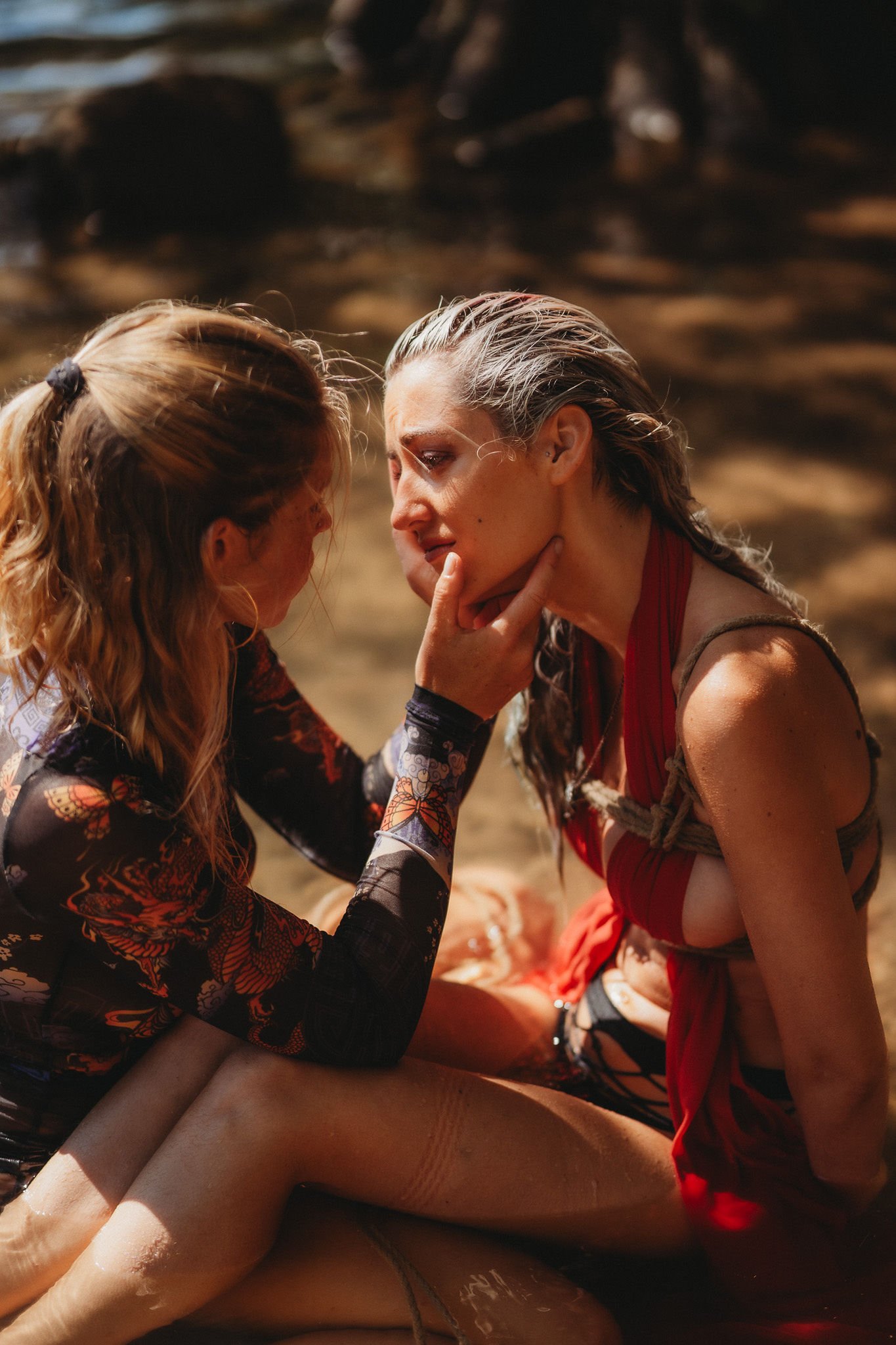 Two women sitting closely near water, one gently touching the other's face, with a comforting and intimate expression, sunlight casting shadows.