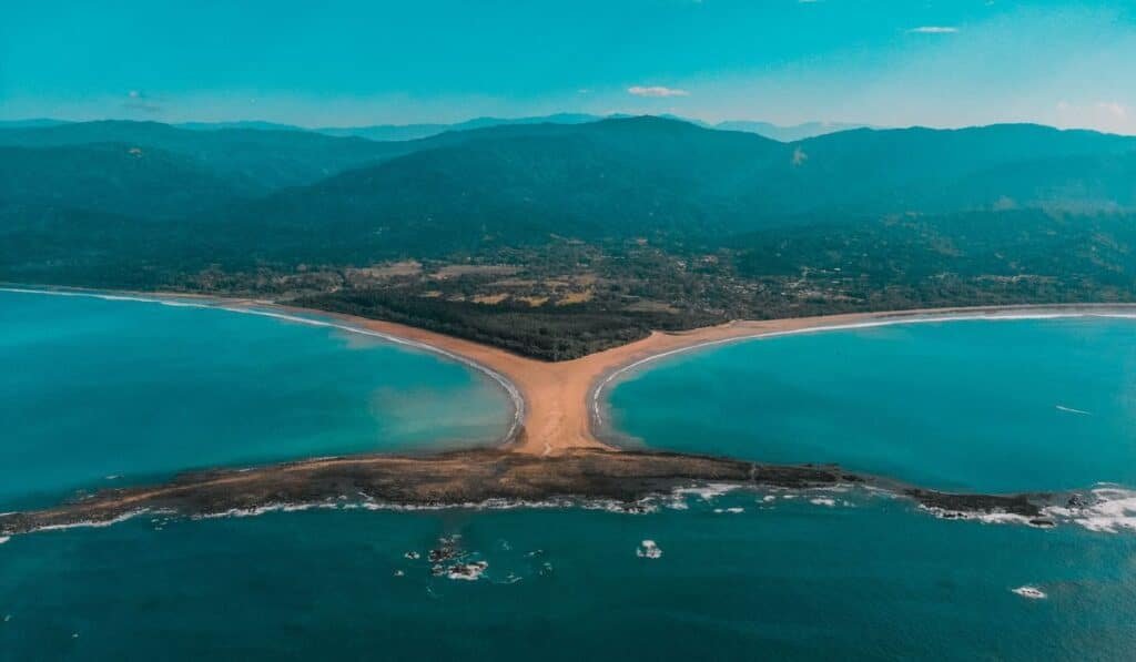 Aerial view of a sandy beach forming a natural whale tail shape with water on both sides, surrounded by lush green hills.