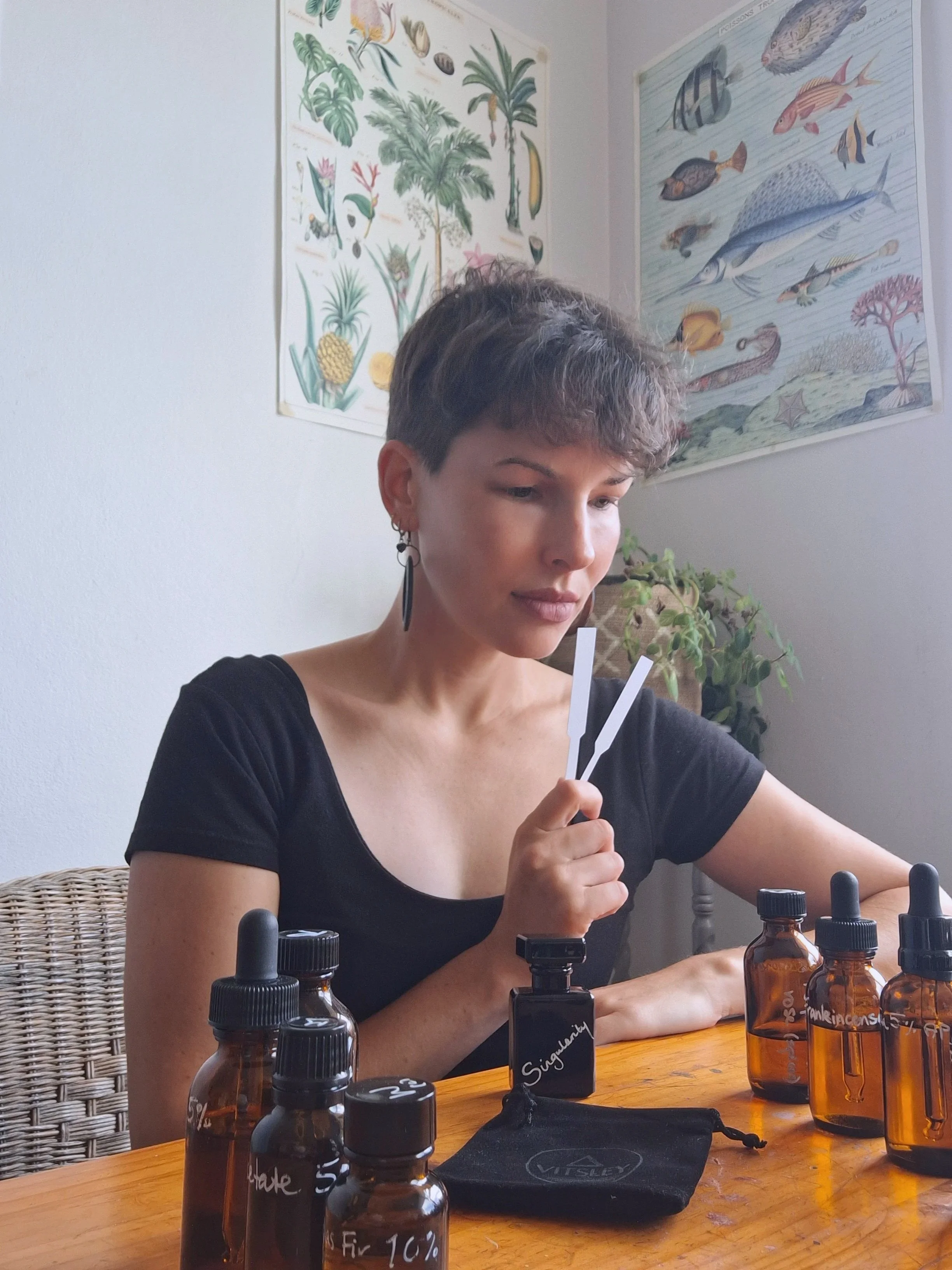 A woman with short, curly hair and earrings sitting at a wooden table with amber bottles labeled with different substances. She appears to be analyzing or working with the bottles, which are likely essential oils or similar products, in a room decorated with botanical and fish posters on the wall.