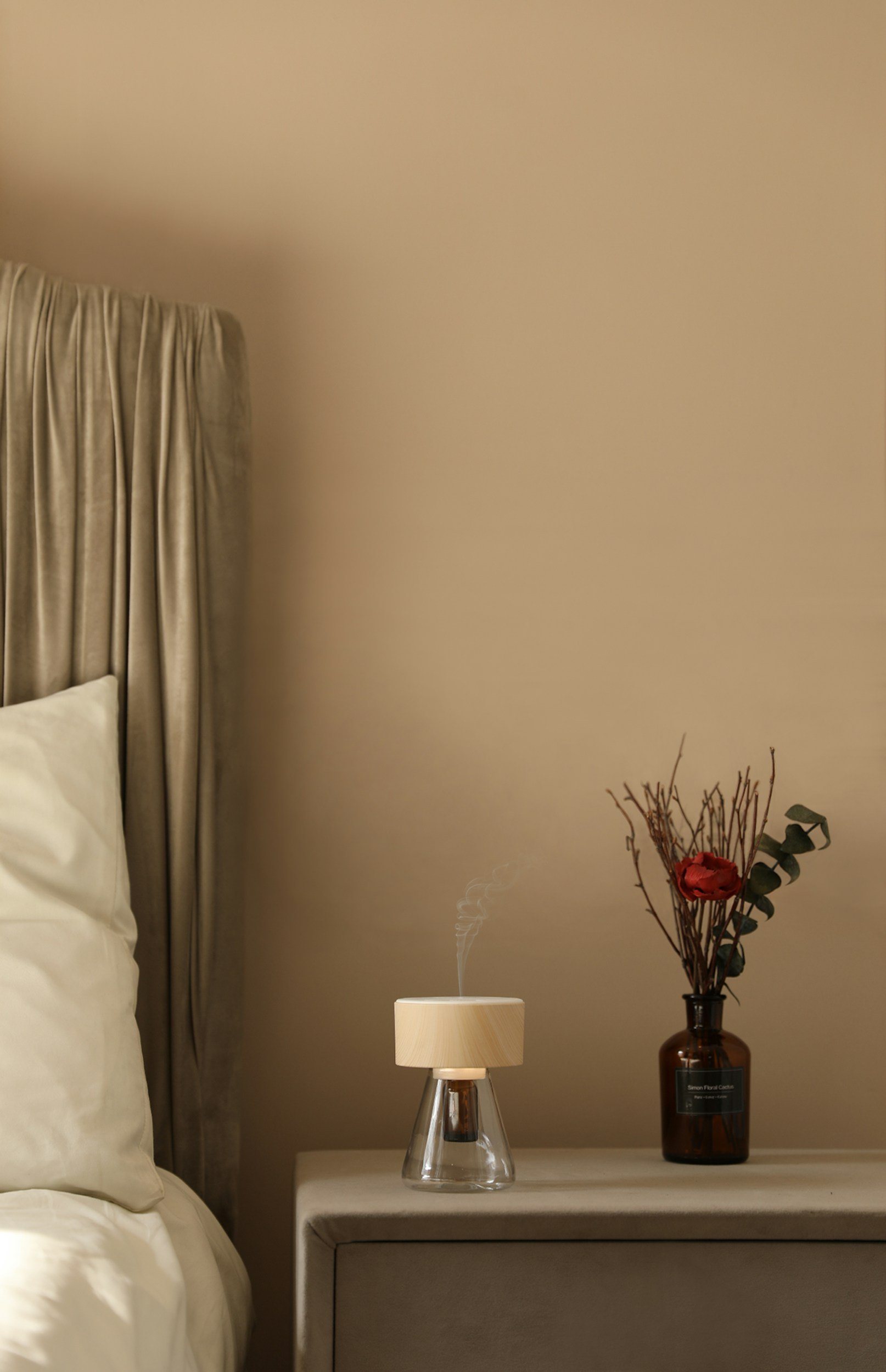 A bedside table with a steam aroma diffuser, a vase with flowers and branches, a beige pillow, and cream-colored curtains.