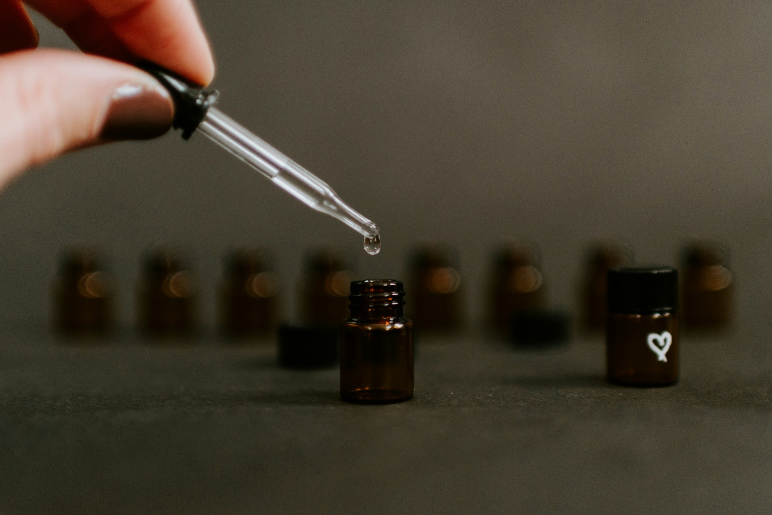 Close-up of a hand holding a glass dropper above small amber bottles, with one bottle in the foreground and several blurred in the background.