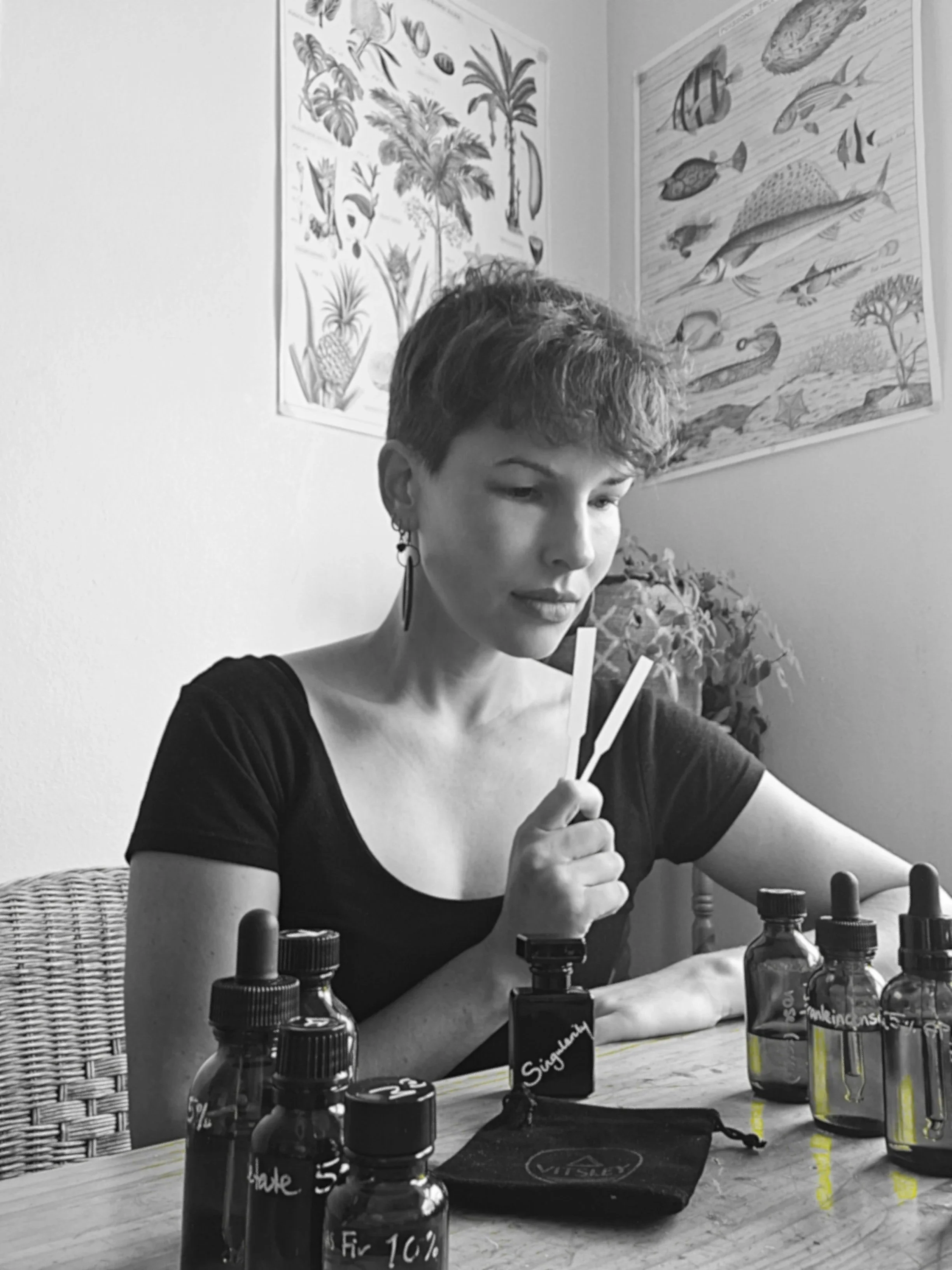 A woman with short hair wearing earrings and a black shirt, sitting at a table with various bottles, some labeled with handwritten notes, and a black pouch, in front of a wall decorated with tropical-themed posters featuring leaves, palm trees, and fish.