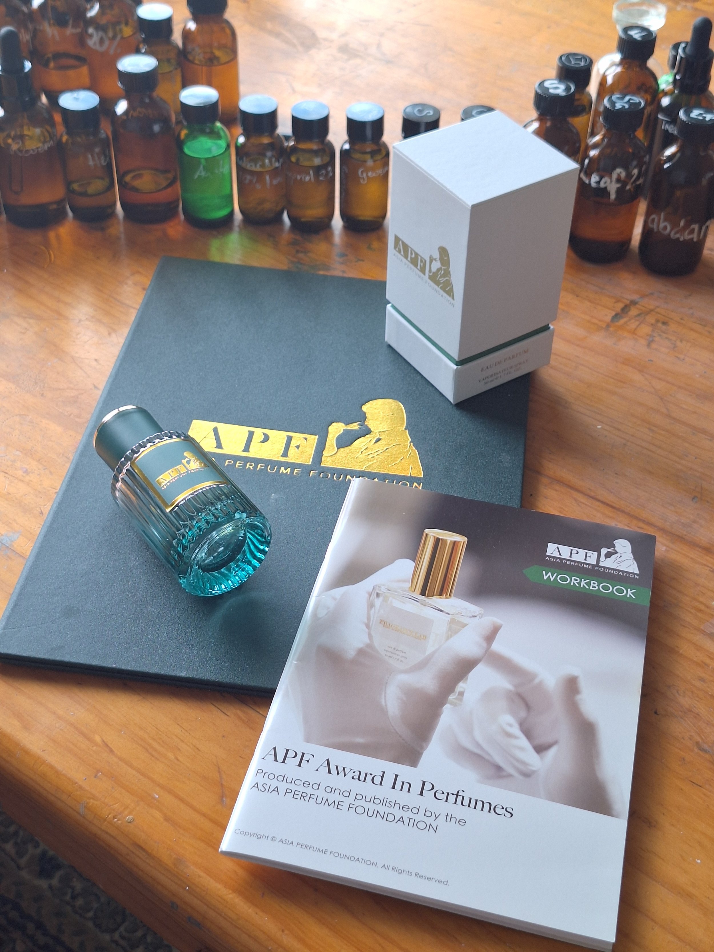 A table with several small bottles of various colors and a box, along with a booklet titled 'APF Award In Perfumes' and a perfume bottle within a hand on the cover.