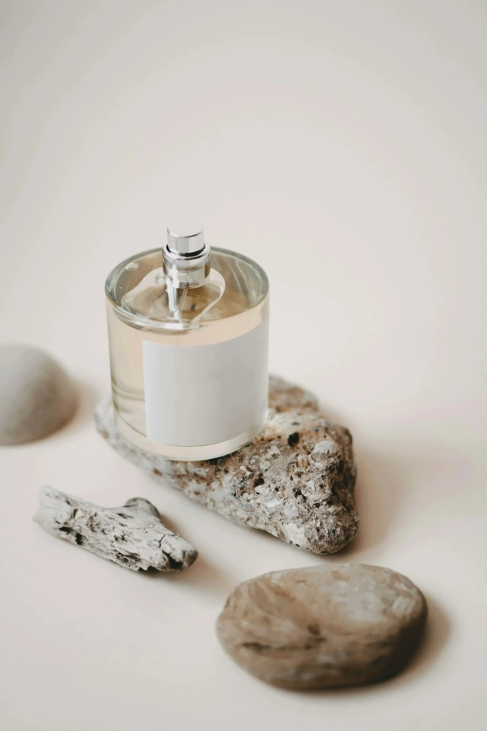 A clear glass perfume bottle with a metallic spray top, placed on a rough granite rock, with pebbles and stones around it on a white surface.
