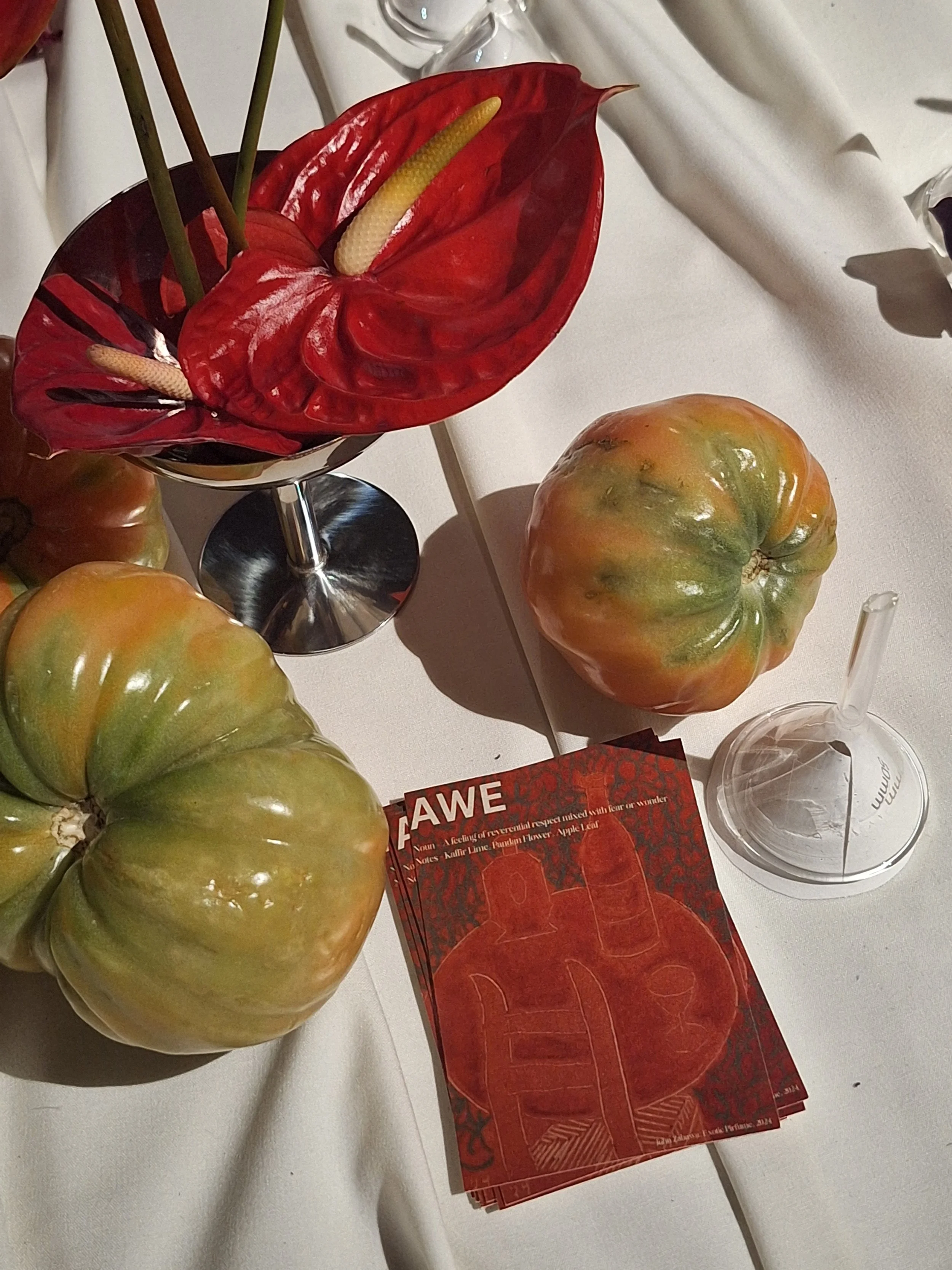 A display of decorative objects on a table, including a greenish tomato, red and yellow leafed flowers, a clear glass or plastic container, a red brochure or flyer with white and black text, and a funnel that has been scented with artistic aroma