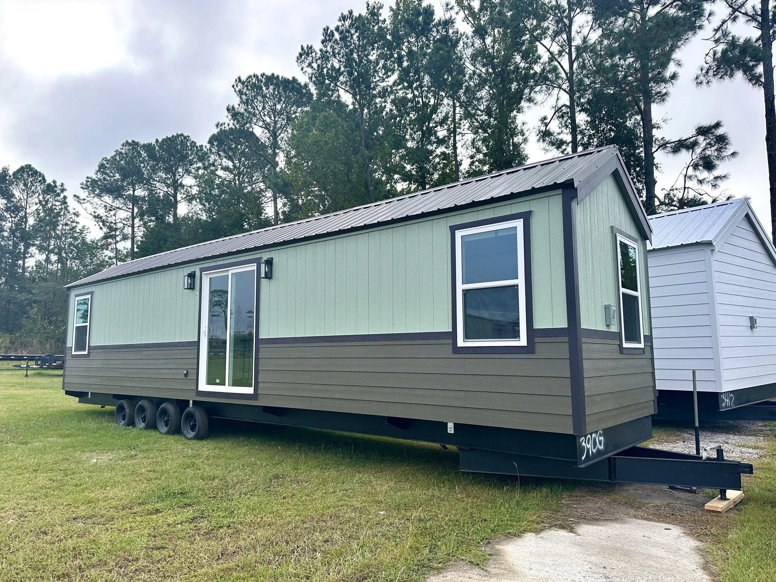 A tiny, modern, green house on wheels with black trim and large windows, situated on a grassy area with tall pine trees in the background under cloudy skies.