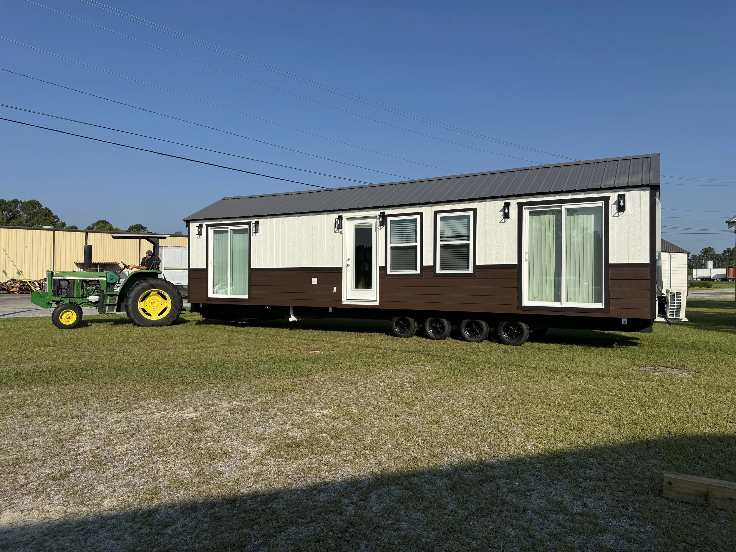 A tiny house on wheels with white and brown panels, multiple windows with curtains, and a gray metal roof. A man is driving a green tractor nearby on a grassy field under a clear blue sky.