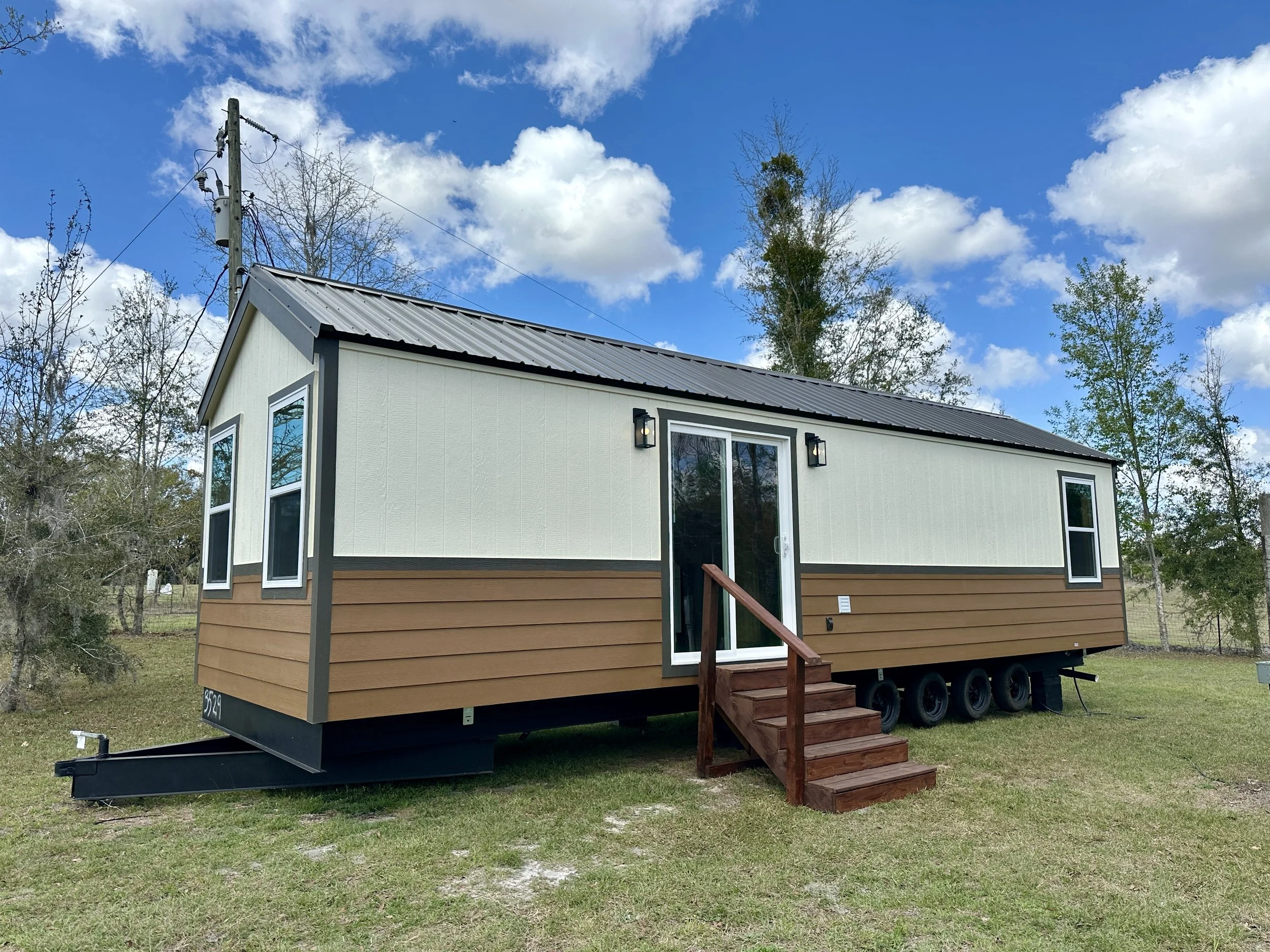 A tiny house on wheels with a cream and light brown exterior, three large windows, a small wooden staircase, and black exterior lights, set outdoors with trees and a partly cloudy sky in the background.