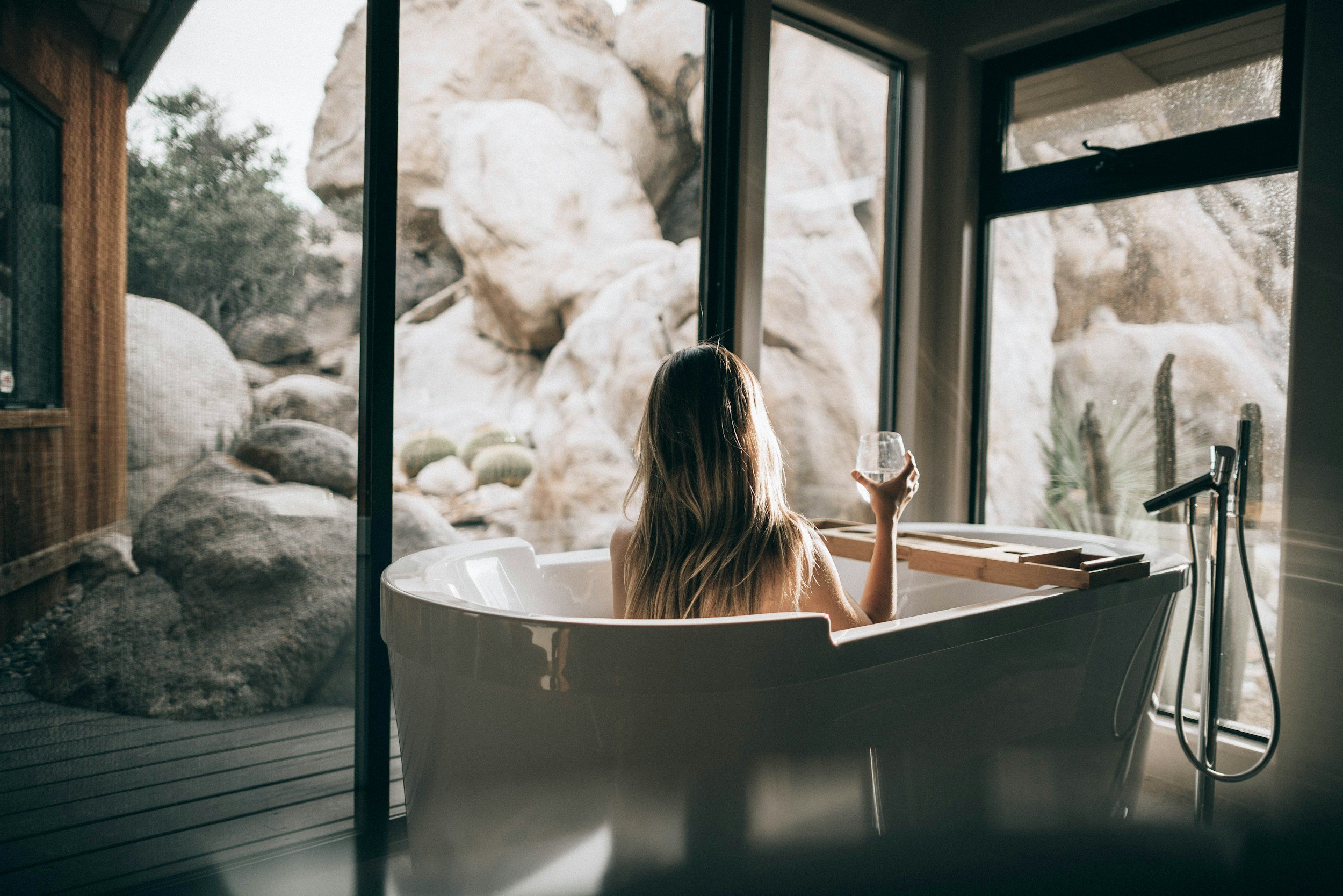 Woman in a bathtub looking out at rocky outdoor scenery through large windows, holding a glass in hand.