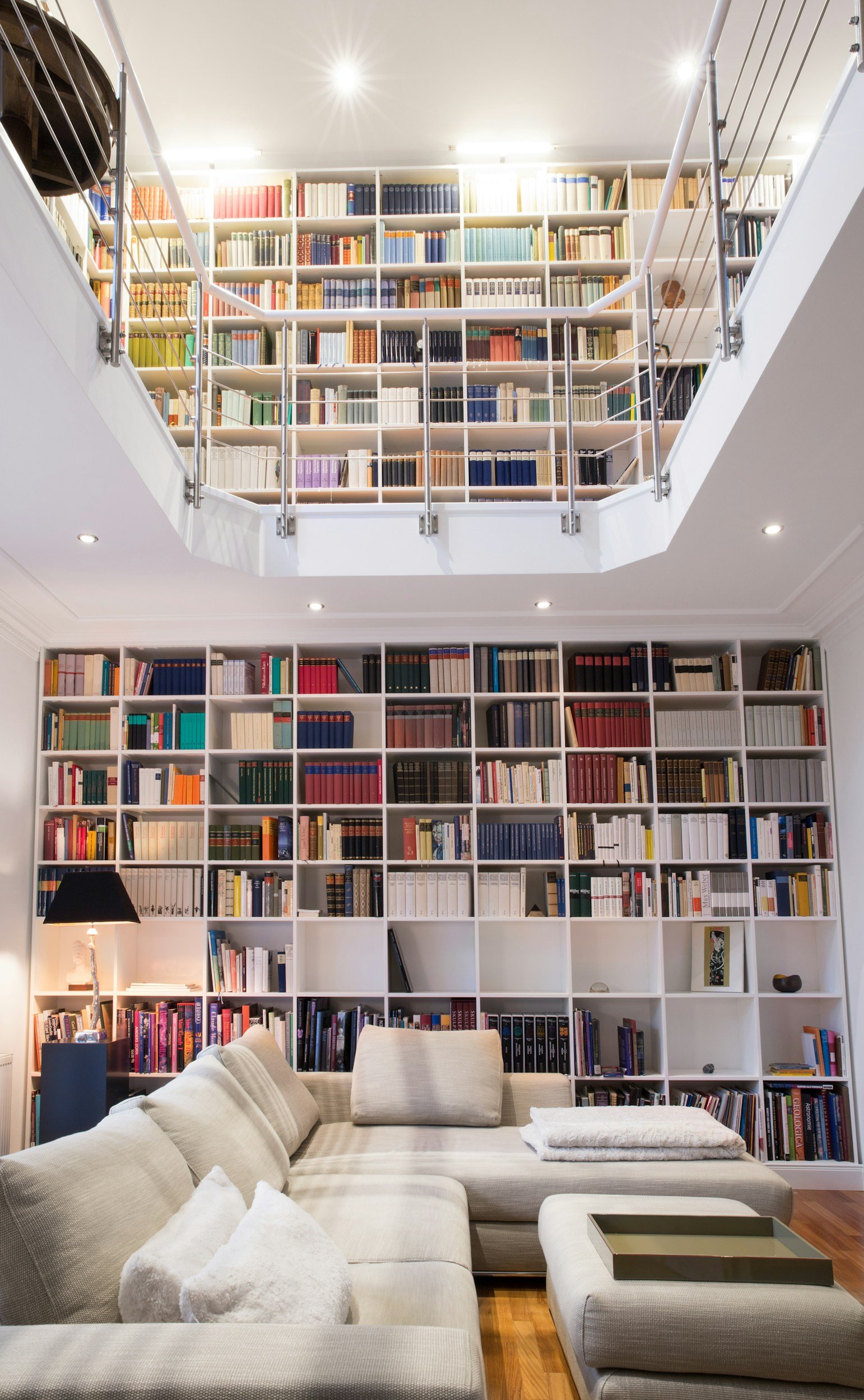 Modern living room with floor-to-ceiling bookshelves filled with colorful books, a gray sectional sofa, a floor lamp, and a wooden floor.