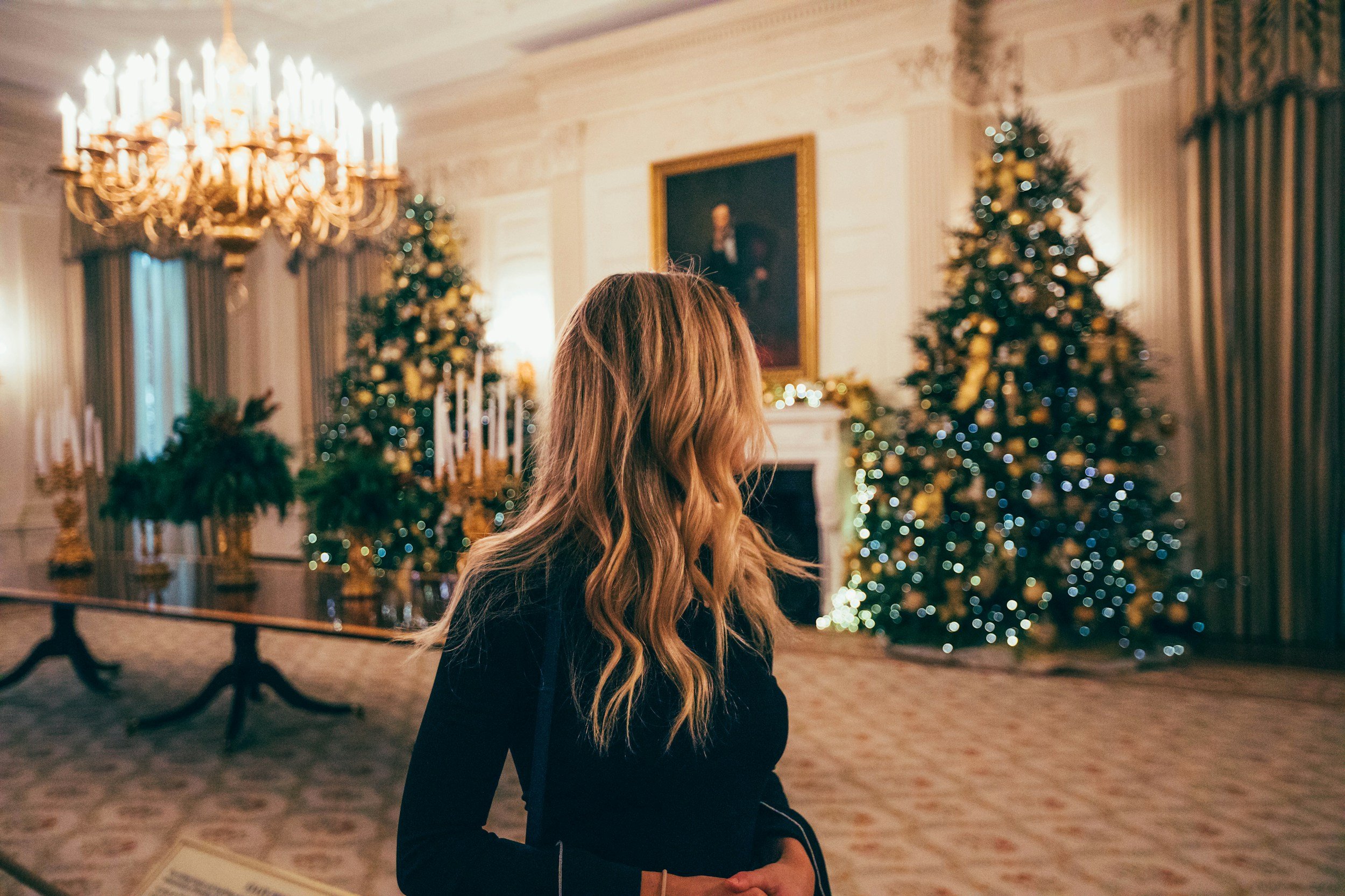 A woman with long hair stands in an elegantly decorated room featuring Christmas trees with lights and ornaments, a chandelier, and candelabras.