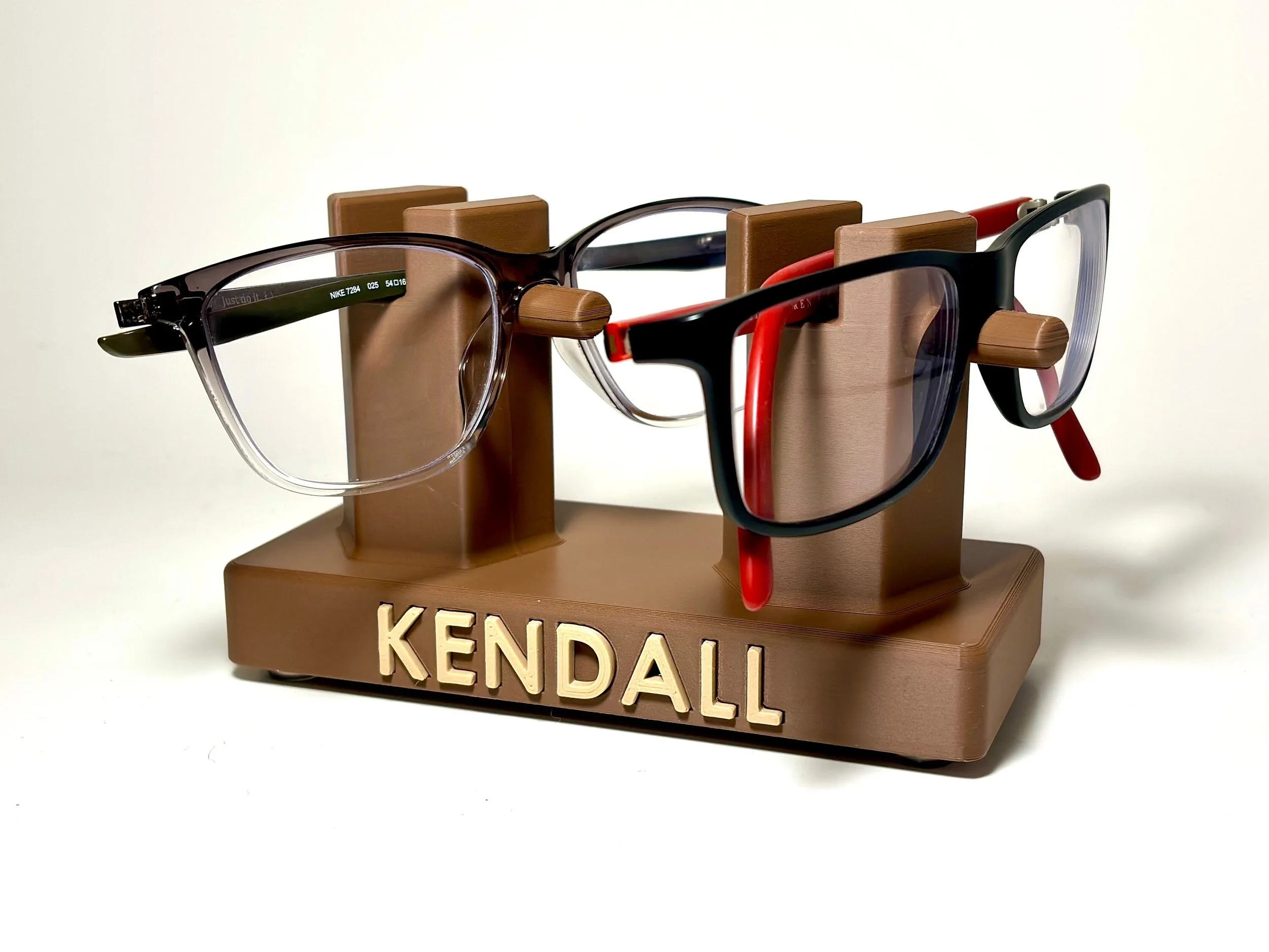 Two pairs of eyeglasses displayed on a brown personalized glasses stand with the word 'KENDALL' in raised letters, one with a black frame and one with a clear and black frame, against a white background.