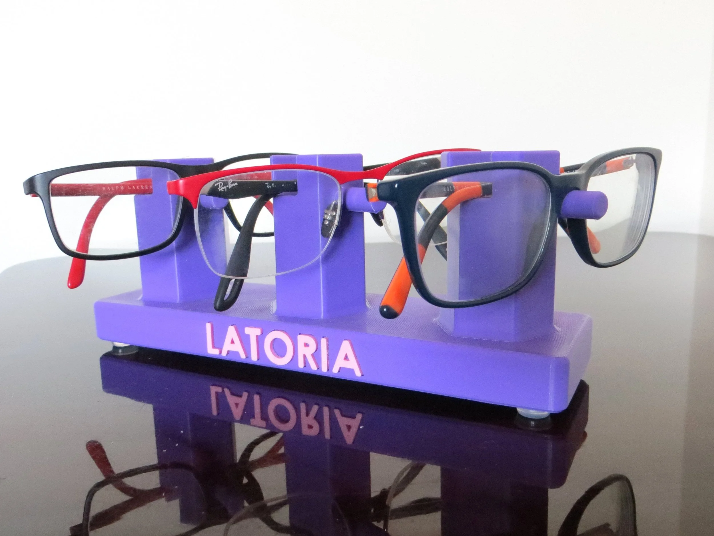Multiple pairs of eyeglasses on a purple personalized glasses display stand labeled 'LATORIA' with reflections on a black surface.