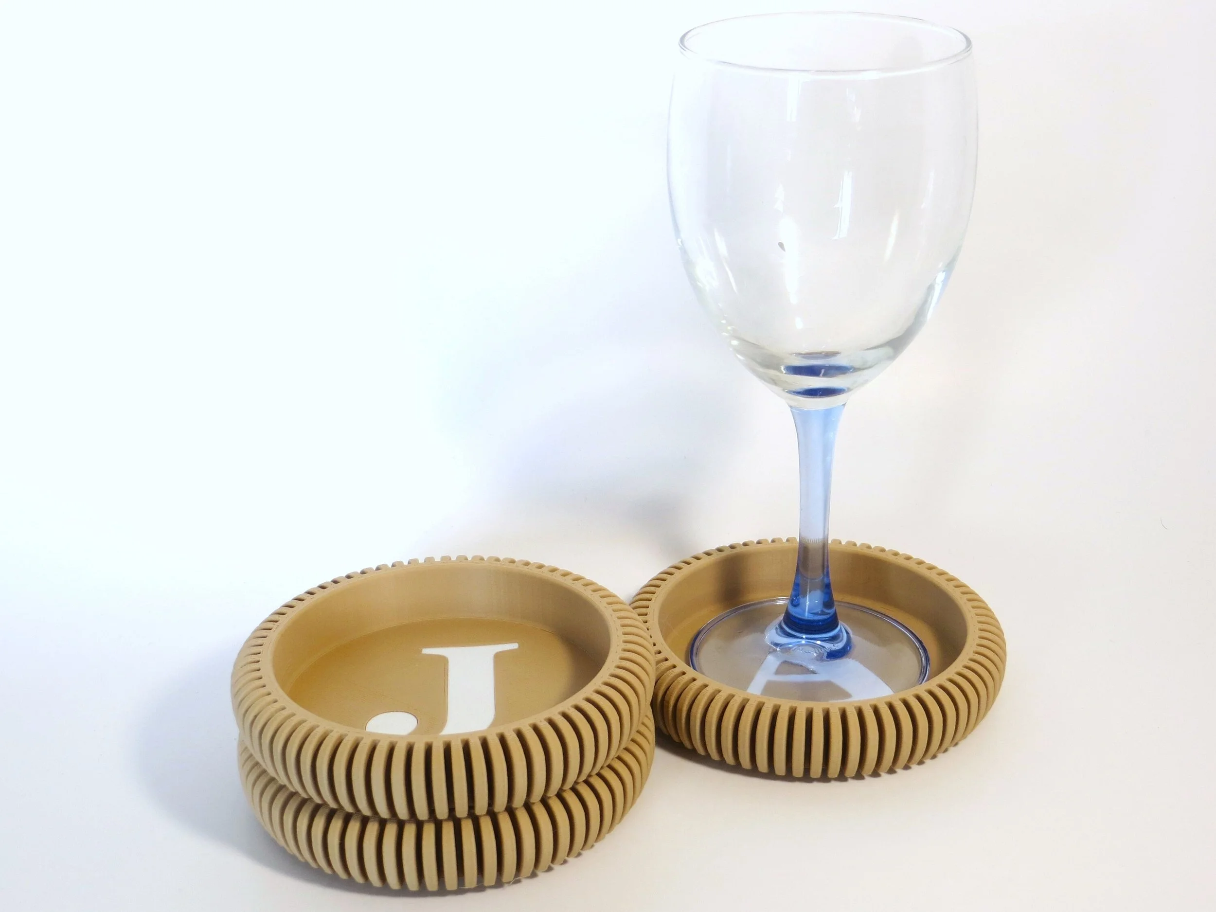A clear wine glass with a blue stem placed on a round cork coaster, with four additional modern coasters stacked beside it, each with a letter printed inside; the visible letter is 'J'.