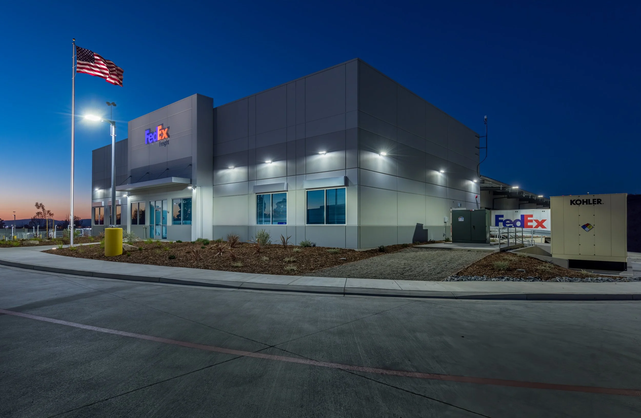 Night view of a FedEx Freight building with an American flag flying in front, illuminated exterior lights, a KOHLER generator, and a clear sky.