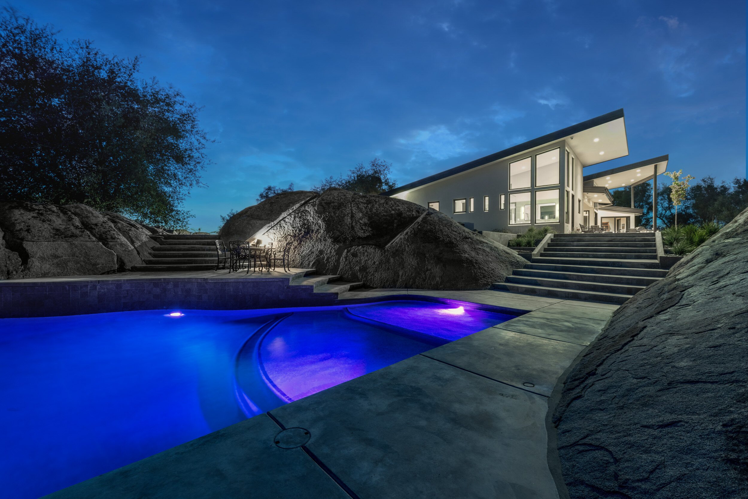 Modern house with large windows and unique sloped roof designs, situated on a hill with stairs leading up to it. The house is lit from within, creating a warm glow against the evening sky. There is a patio area with outdoor furniture and a small tree
