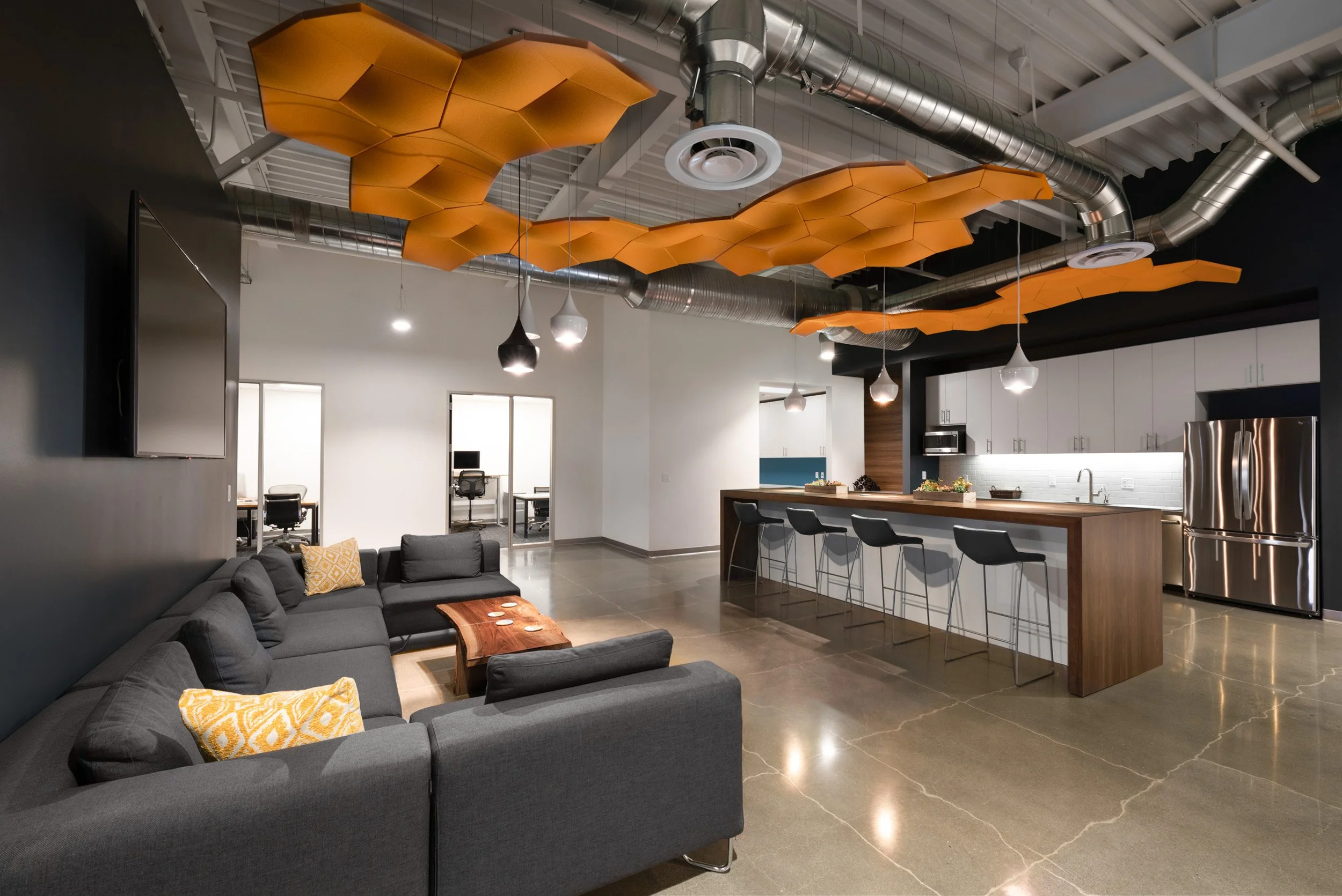 Modern open-concept office lounge and kitchen area with gray sectional sofa, geometric orange ceiling acoustic panels, black pendant lighting, stainless steel ventilation ducts, white and wood kitchen cabinets, bar seating, and a refrigerator.