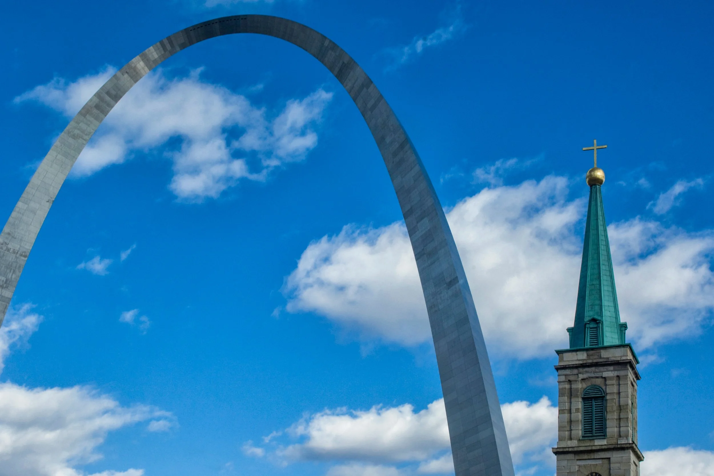 The Gateway Arch in St. Louis, Missouri, with a church steeple topped with a cross in the foreground, against a partly cloudy blue sky.