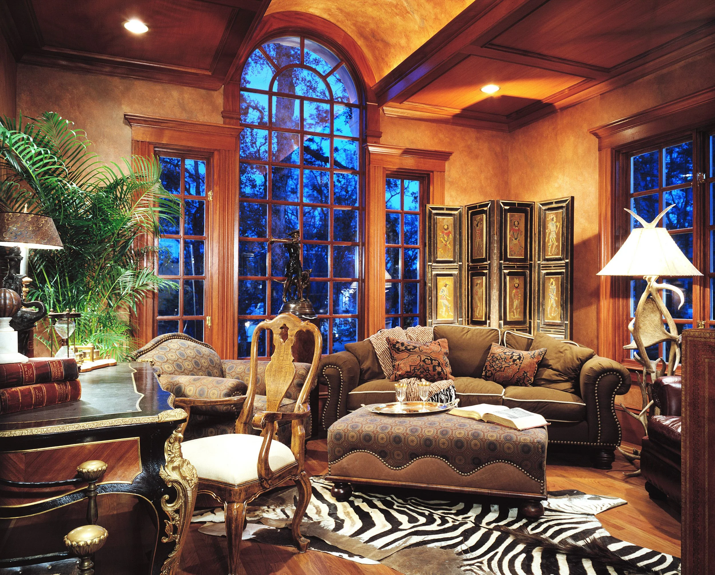 Elegant living room with large arched windows showing blue evening sky and trees outside. The room features wooden crown molding, a zebra print rug, comfortable sofas with patterned pillows, a decorative folding room divider, a table with books, lamp
