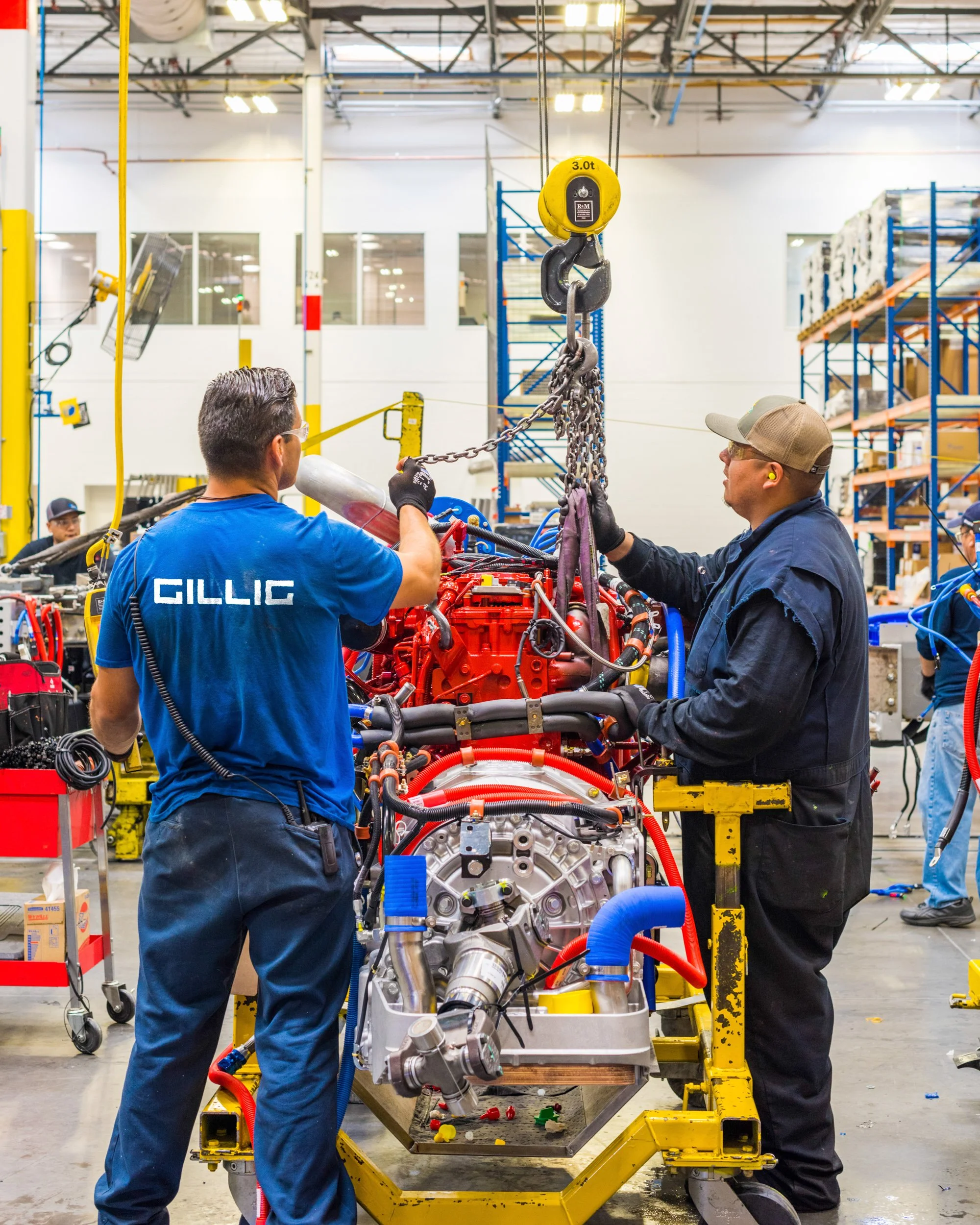 Two workers in a factory working on an engine or large mechanical component mounted on a yellow work platform. One worker is wearing a blue shirt with the name "GILLIC" on the back, and the other is in a dark jacket and beige cap. The background show