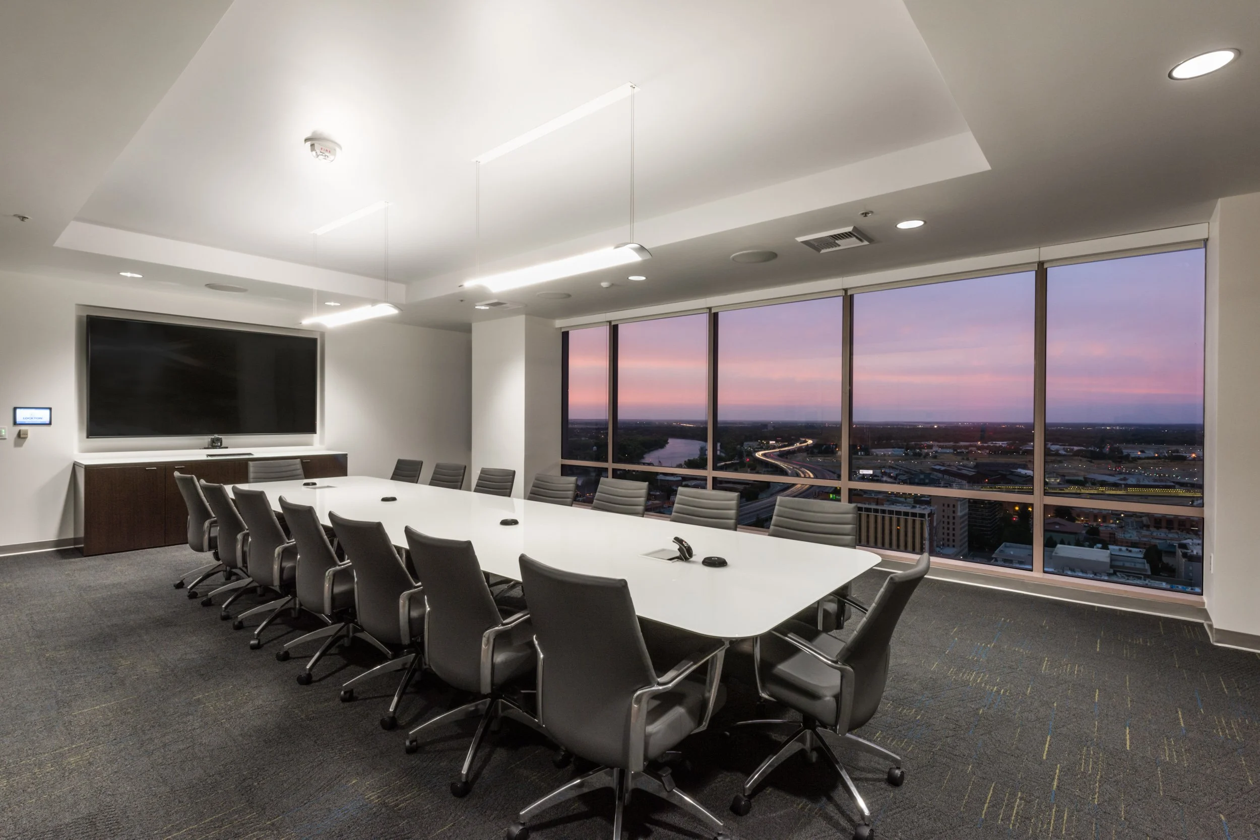 A modern conference room with a white table surrounded by gray chairs, large windows showing a cityscape at sunset, and ceiling lights.