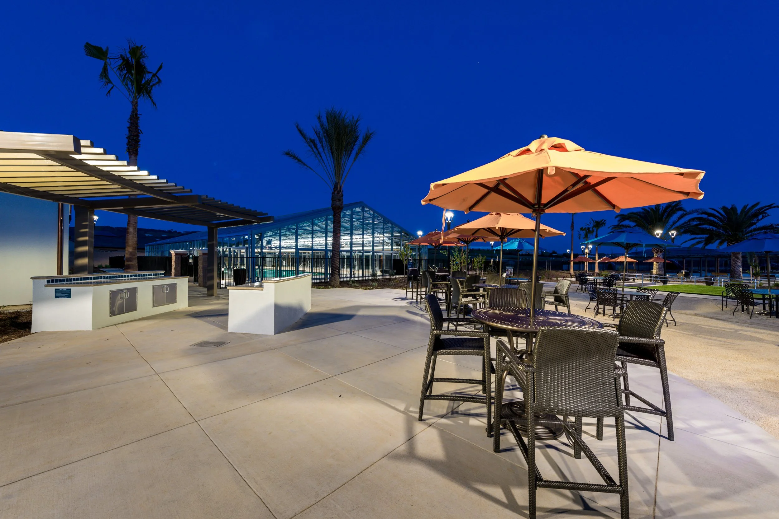 Outdoor seating area with tables, chairs, large umbrellas, palm trees, and a glass greenhouse in the background at dusk.