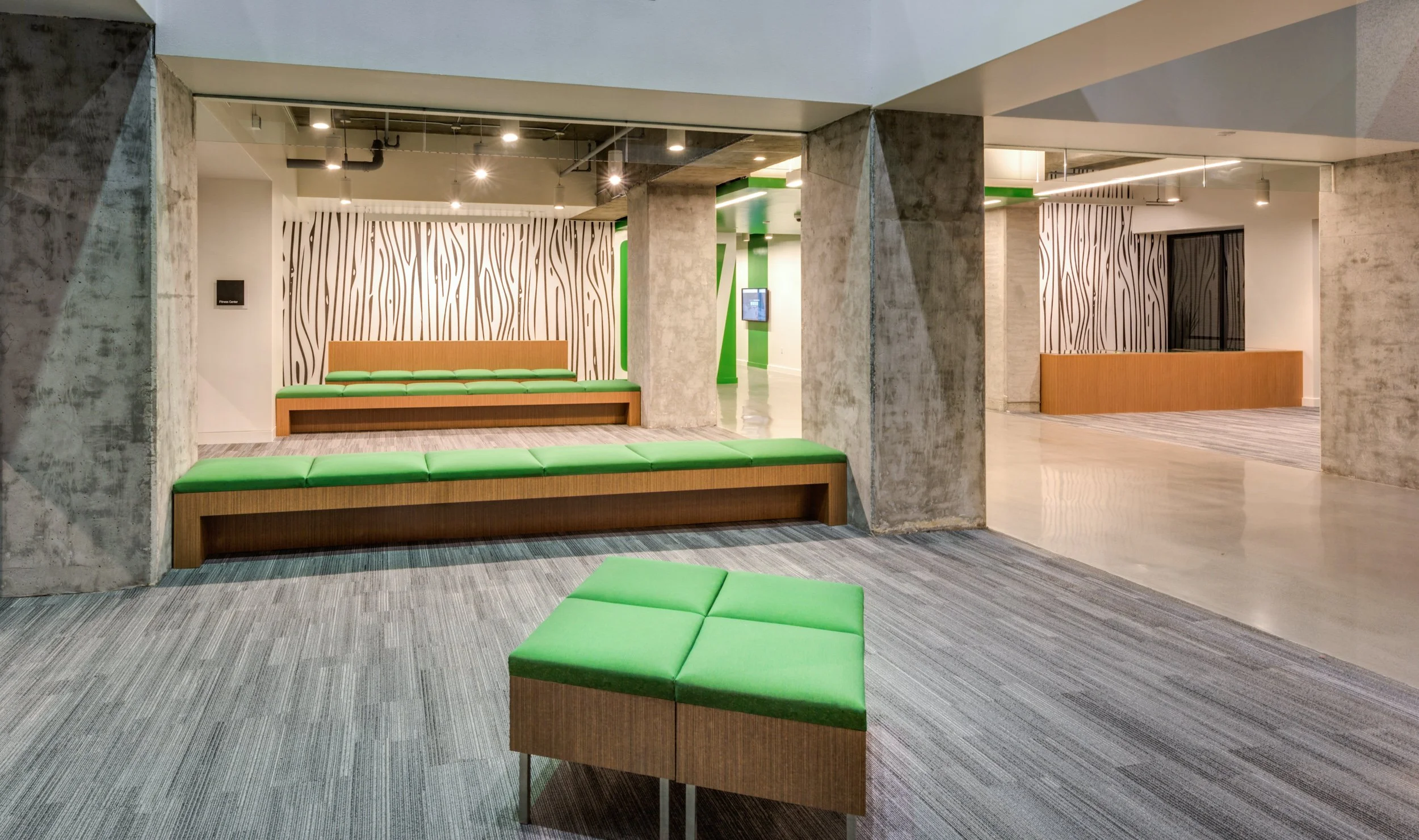 Modern office lobby with concrete columns, green cushioned benches, wooden furniture, patterned wall panels, and a gray carpet.