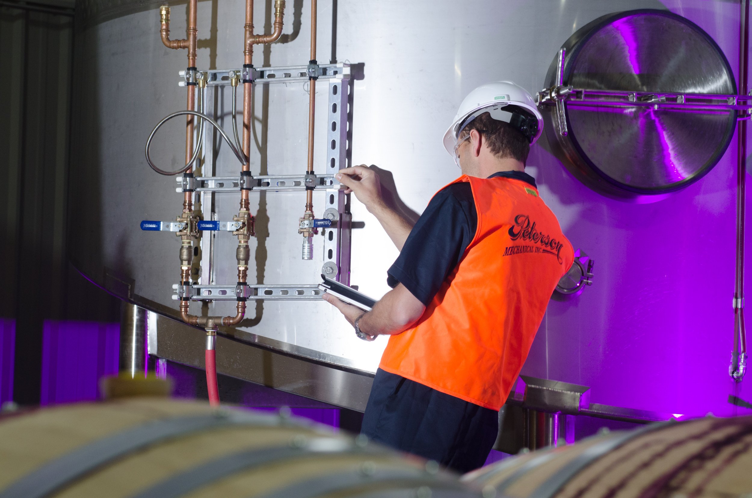 A worker in safety gear, including a hard hat and orange vest, inspecting control panel with copper pipes and valves beside a large industrial tank with purple lighting.