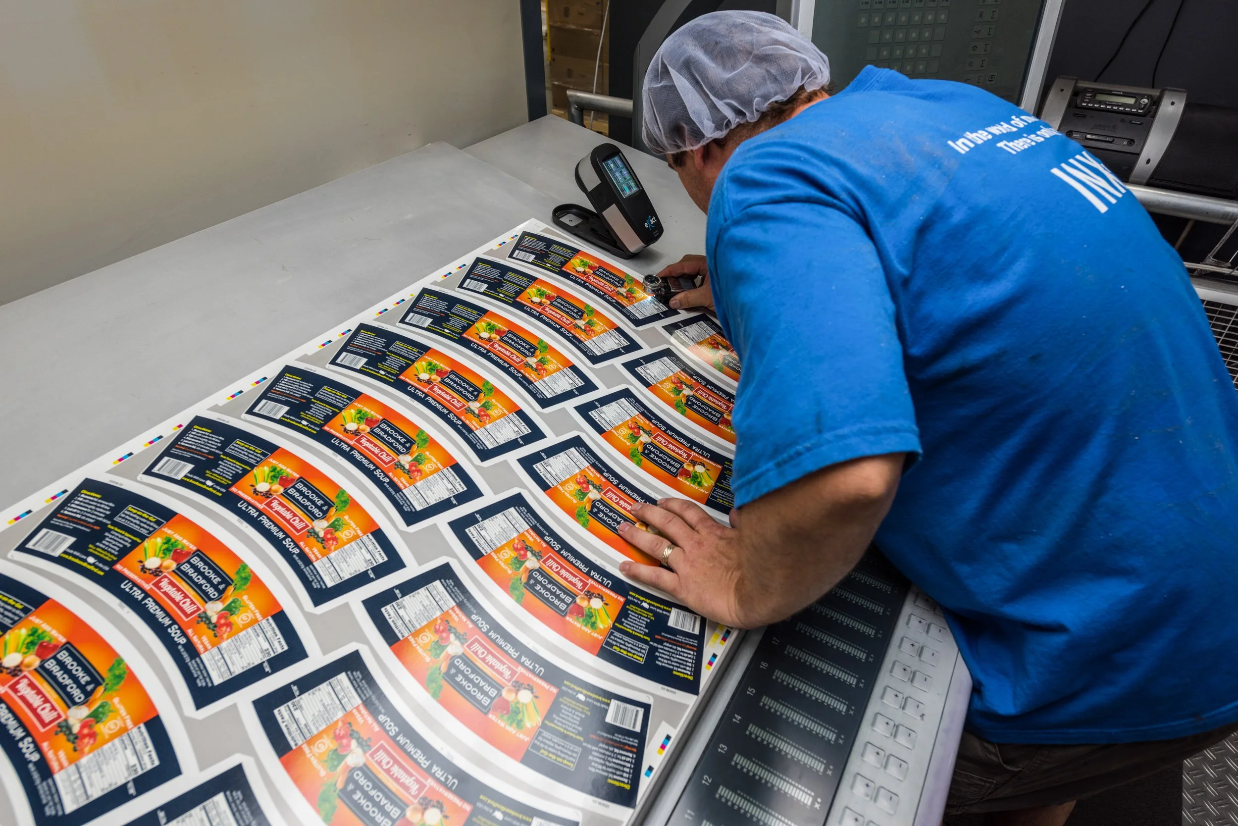 Worker in a blue shirt and hairnet inspecting printed labels on a production line with a computer and printing equipment nearby.