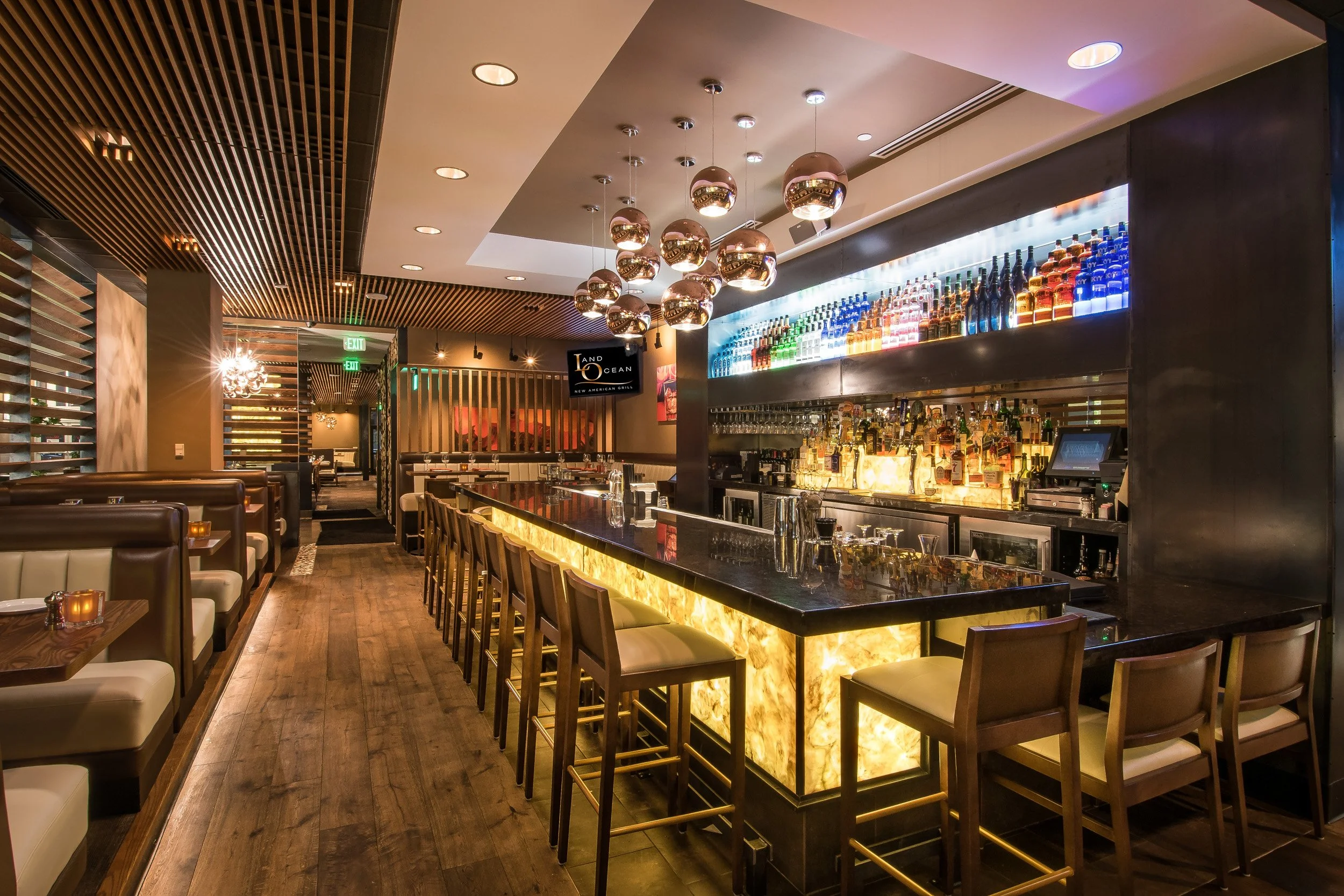 Modern restaurant interior featuring a sleek bar with illuminated onyx countertop, bar stools, and a display of liquor bottles on a backlit shelf. Warm lighting, wood accents, and contemporary decor create an inviting atmosphere.