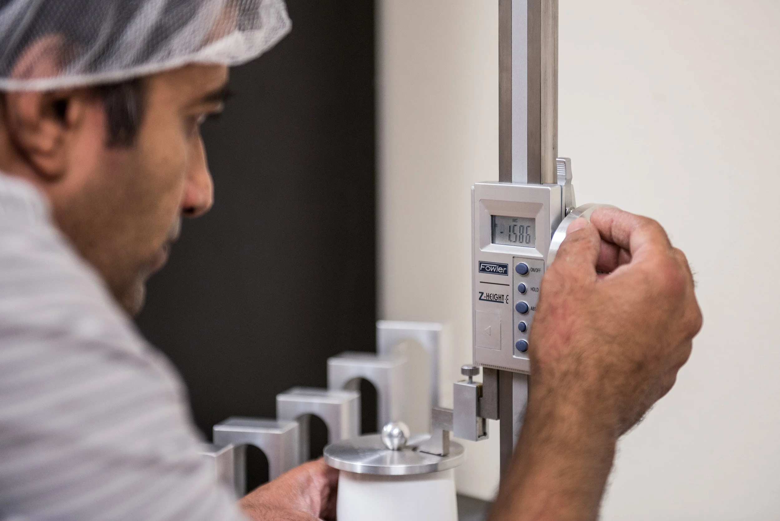 A scientist or engineer in a lab coat and hair cap using a digital height gauge to measure an object, with the gauge showing a reading of -15.86.