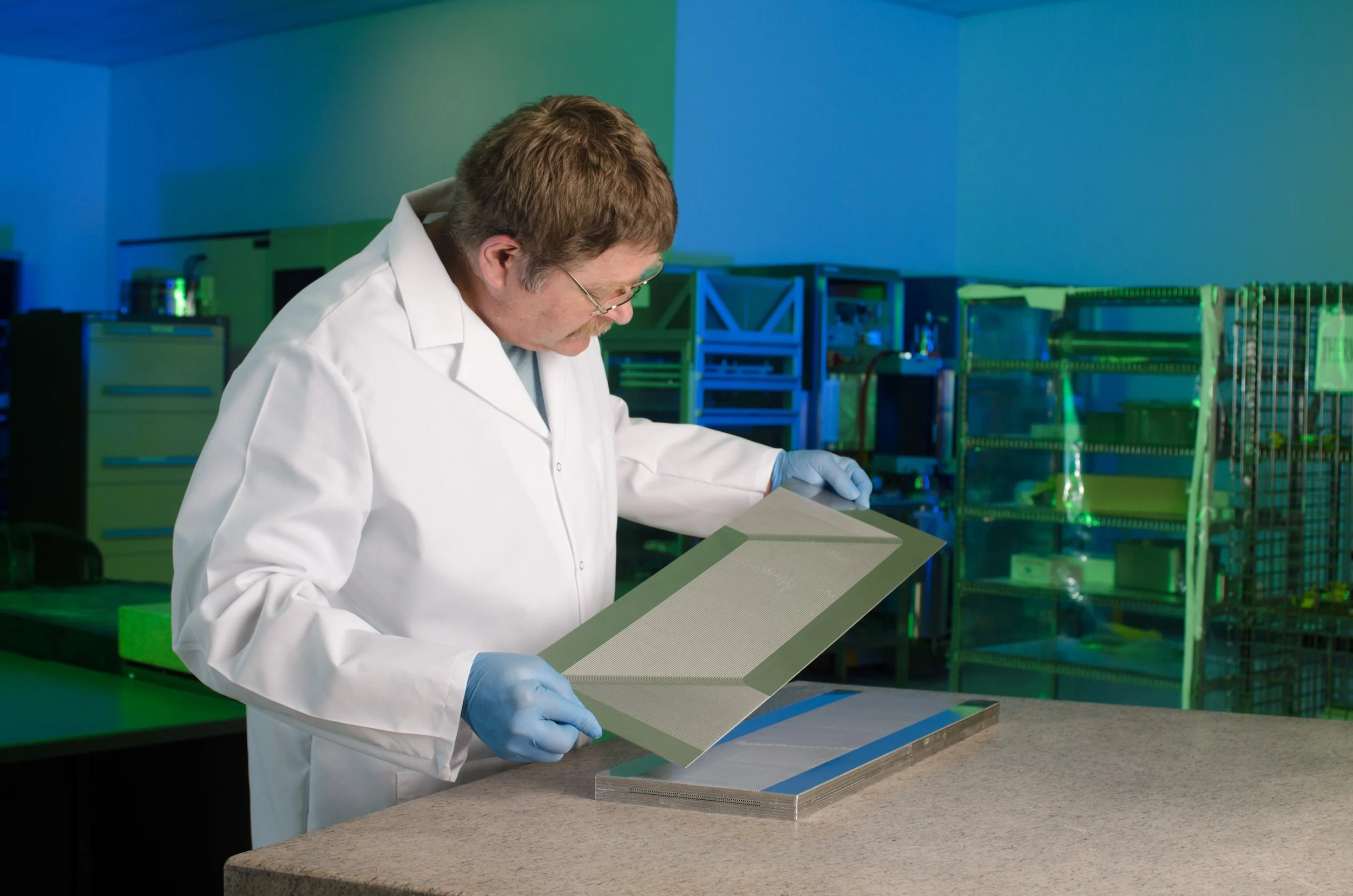 A scientist in a white lab coat and blue gloves examines a scientific sample or material in a laboratory with colorful storage racks and equipment in the background.