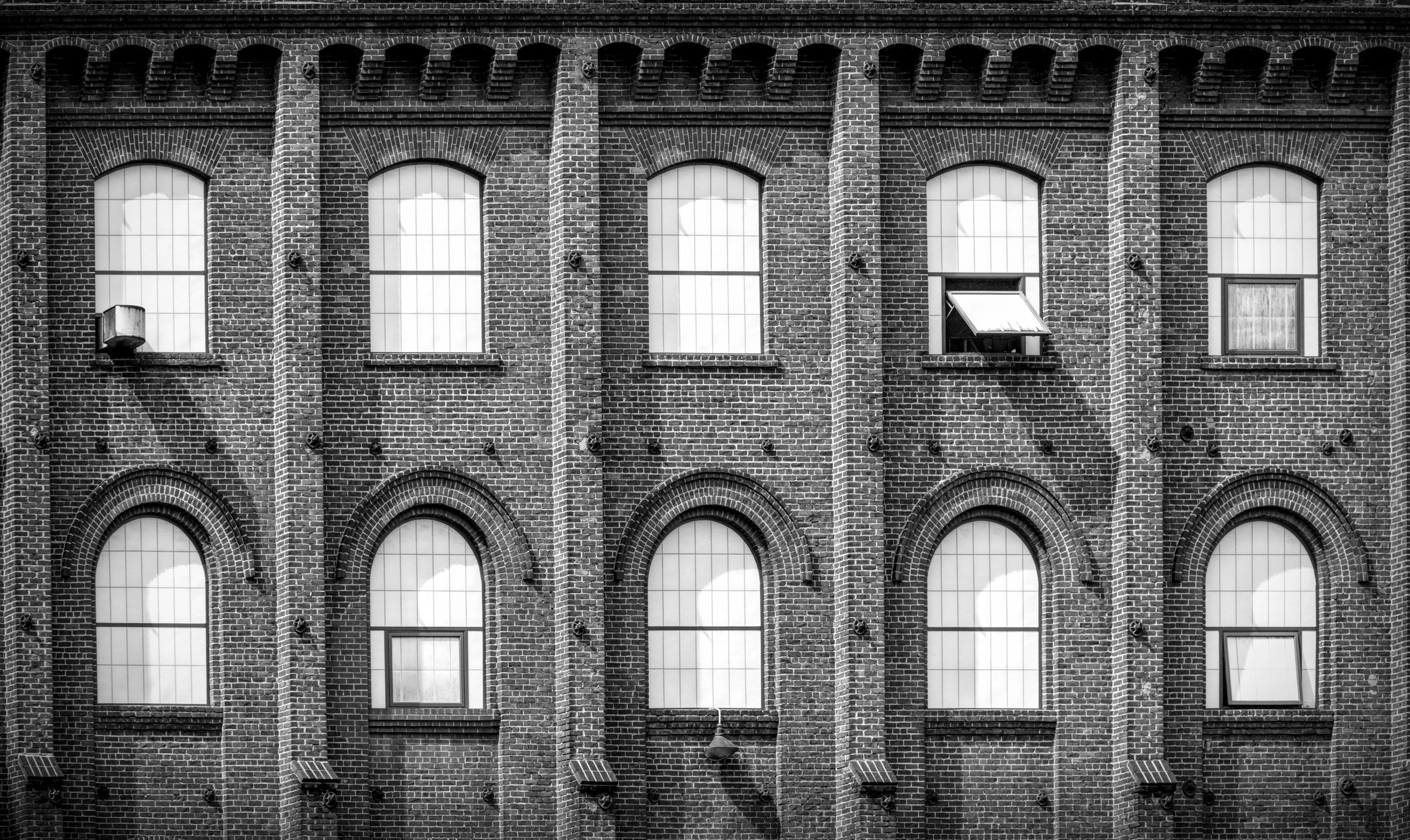 Black and white photo of a brick building's facade with arched windows, some open and some with curtains.