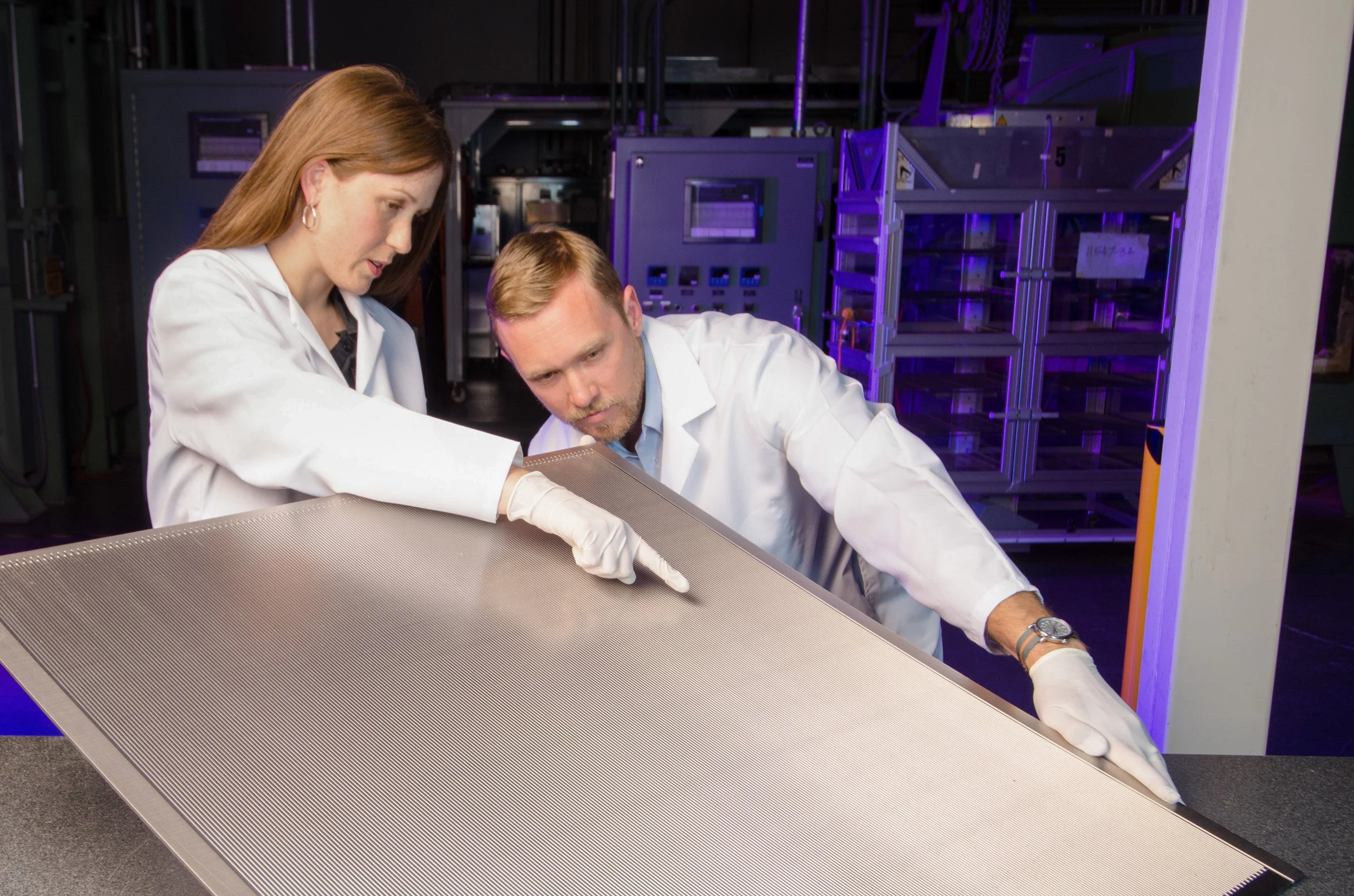 Two scientists in lab coats examining a large metallic material sample in a laboratory with electronic equipment and servers in the background.