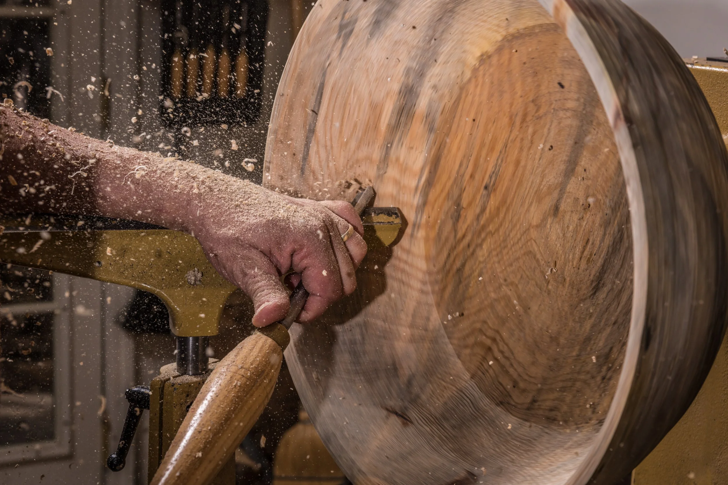 A person is woodworking, spinning a large wooden bowl on a lathe, using a tool to carve and shape the wood, with wood shavings flying in the air.