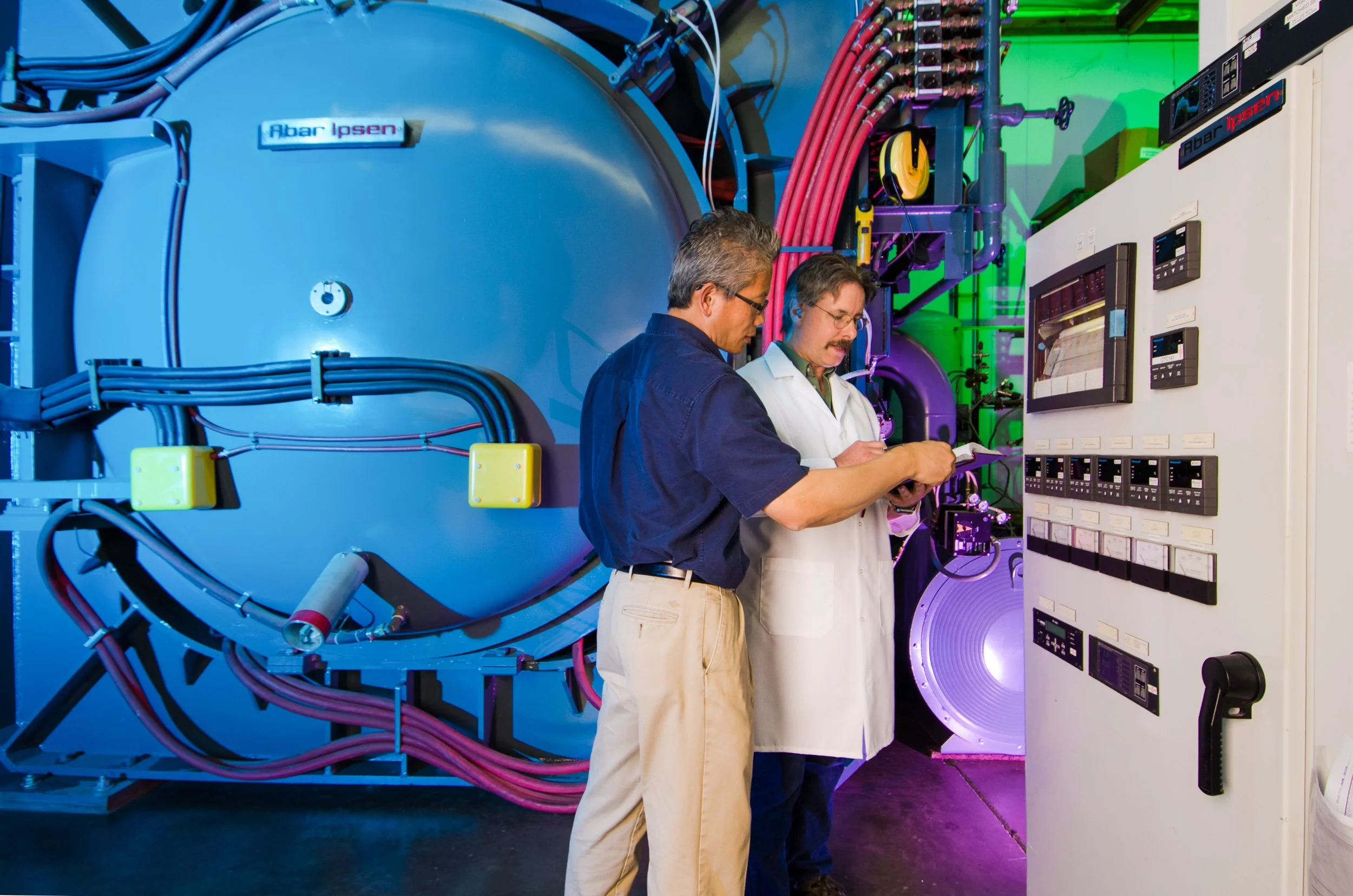 Two men working together in a laboratory setting, inspecting equipment with control panels and large machinery in the background.