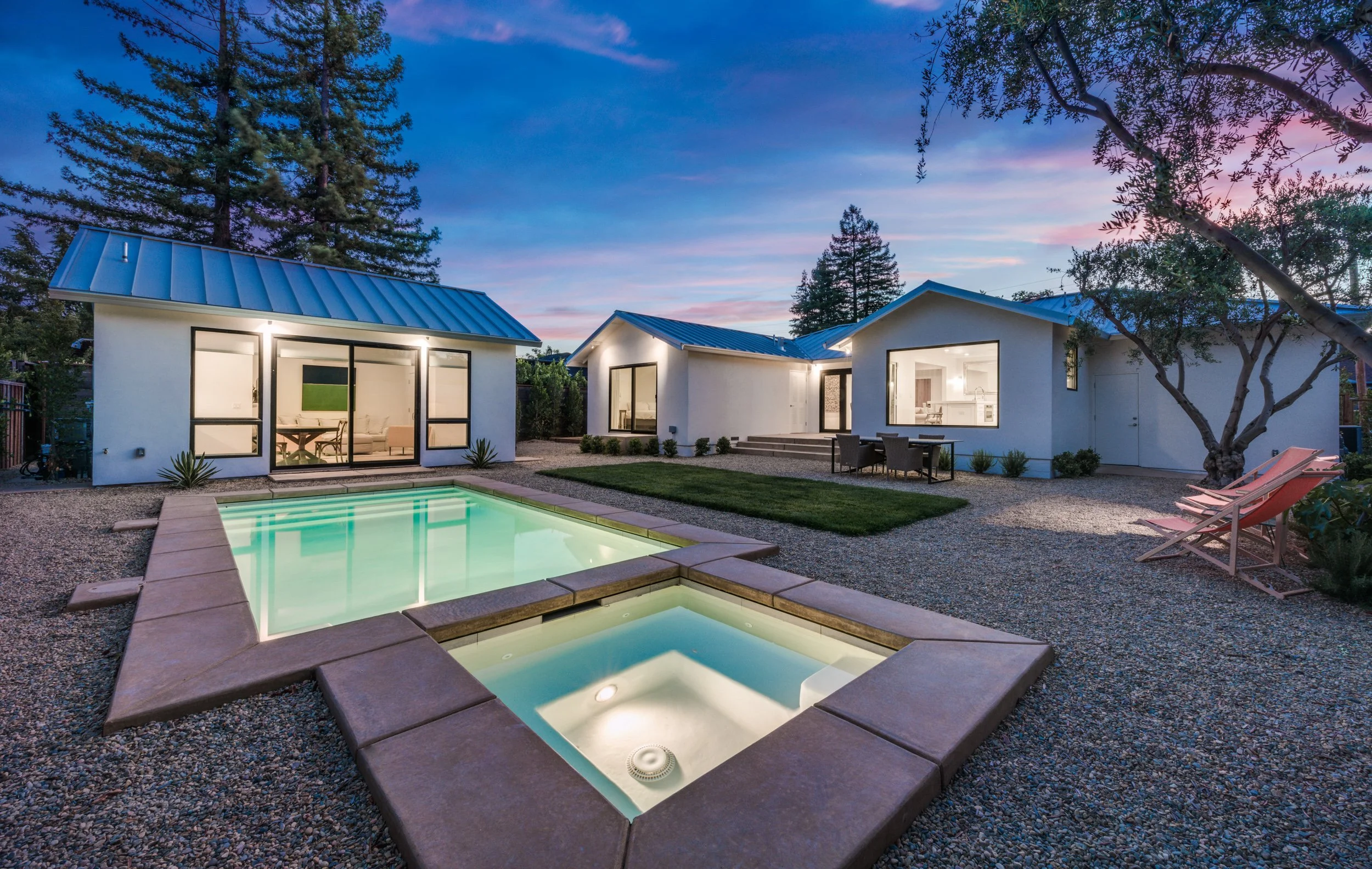 A modern backyard at dusk with a swimming pool, two small white houses with metal roofs, outdoor furniture, and tall trees in the background, illuminated by soft outdoor lighting.