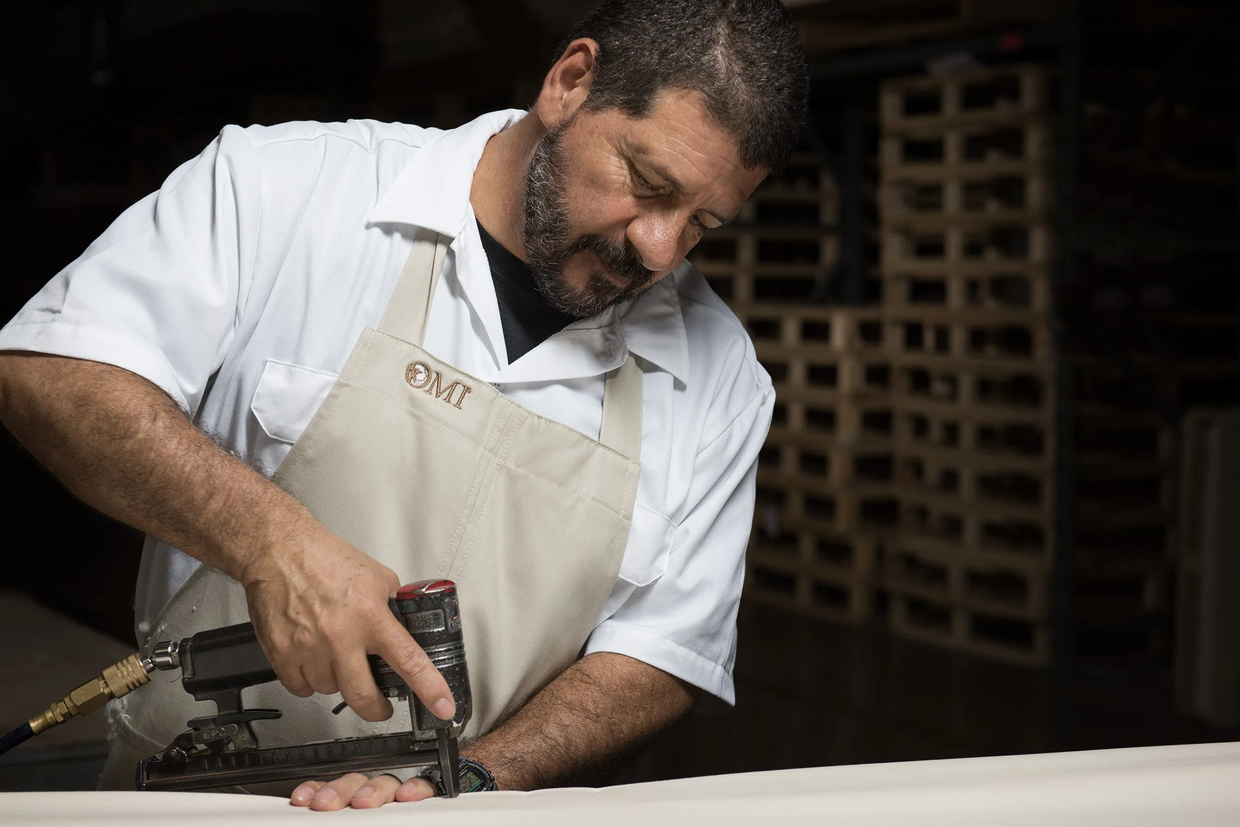 A man in a white apron and black shirt, with a beard and short hair, is using a pneumatic stapler to attach fabric to a surface in a workshop or industrial setting.