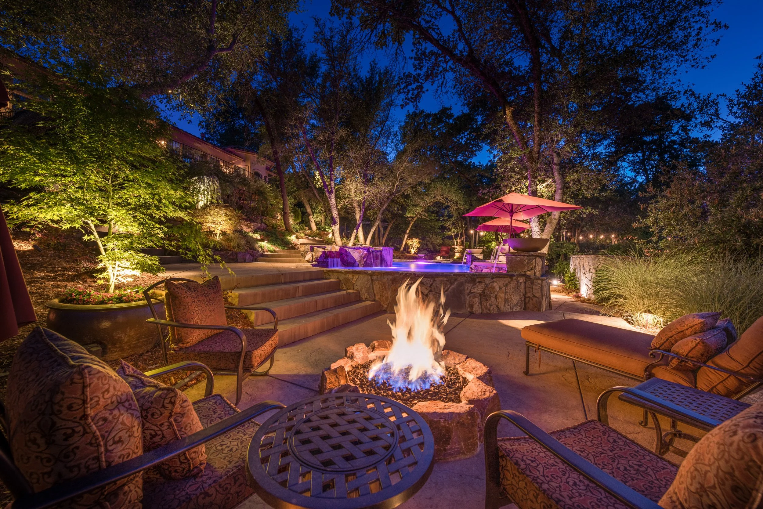 Nighttime outdoor backyard scene with patio furniture, a fire pit, trees, and lit umbrellas.