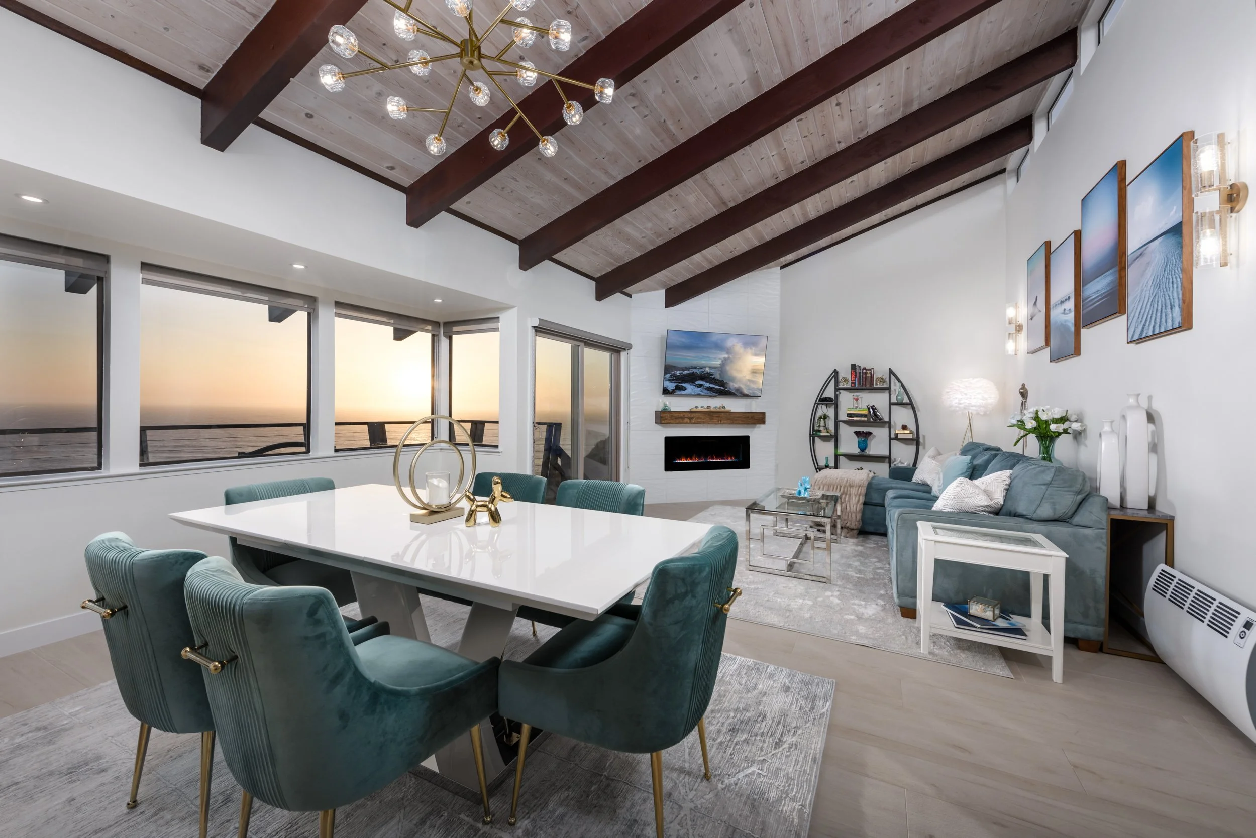 A modern living room with a high wooden ceiling featuring exposed dark beams, a gold chandelier, a flat-screen TV above a fireplace, large windows with a sunset view, a blue sectional sofa, a white coffee table, decorative vases, and framed wall art.