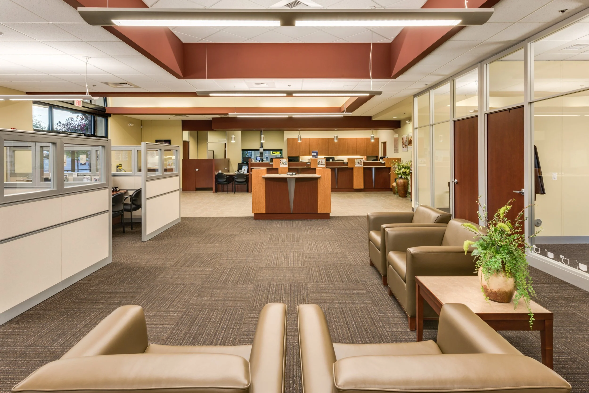 Empty waiting room with brown and beige furniture, a reception desk in the background, large windows, and potted plants.