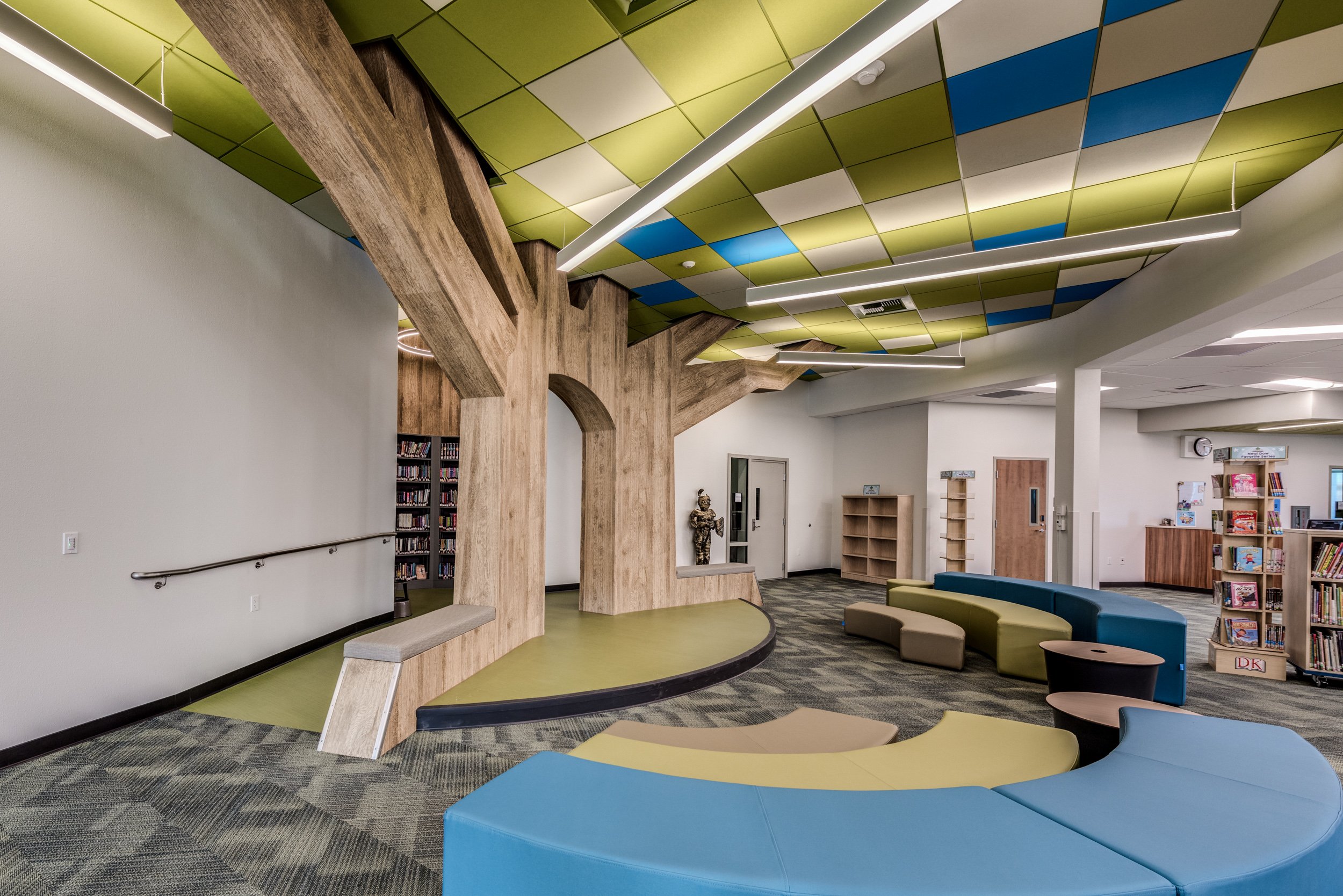 Interior of a modern library or children’s area with curved seating, a wooden tree-shaped structure, bookshelves, and colorful ceiling panels in green, white, and blue.