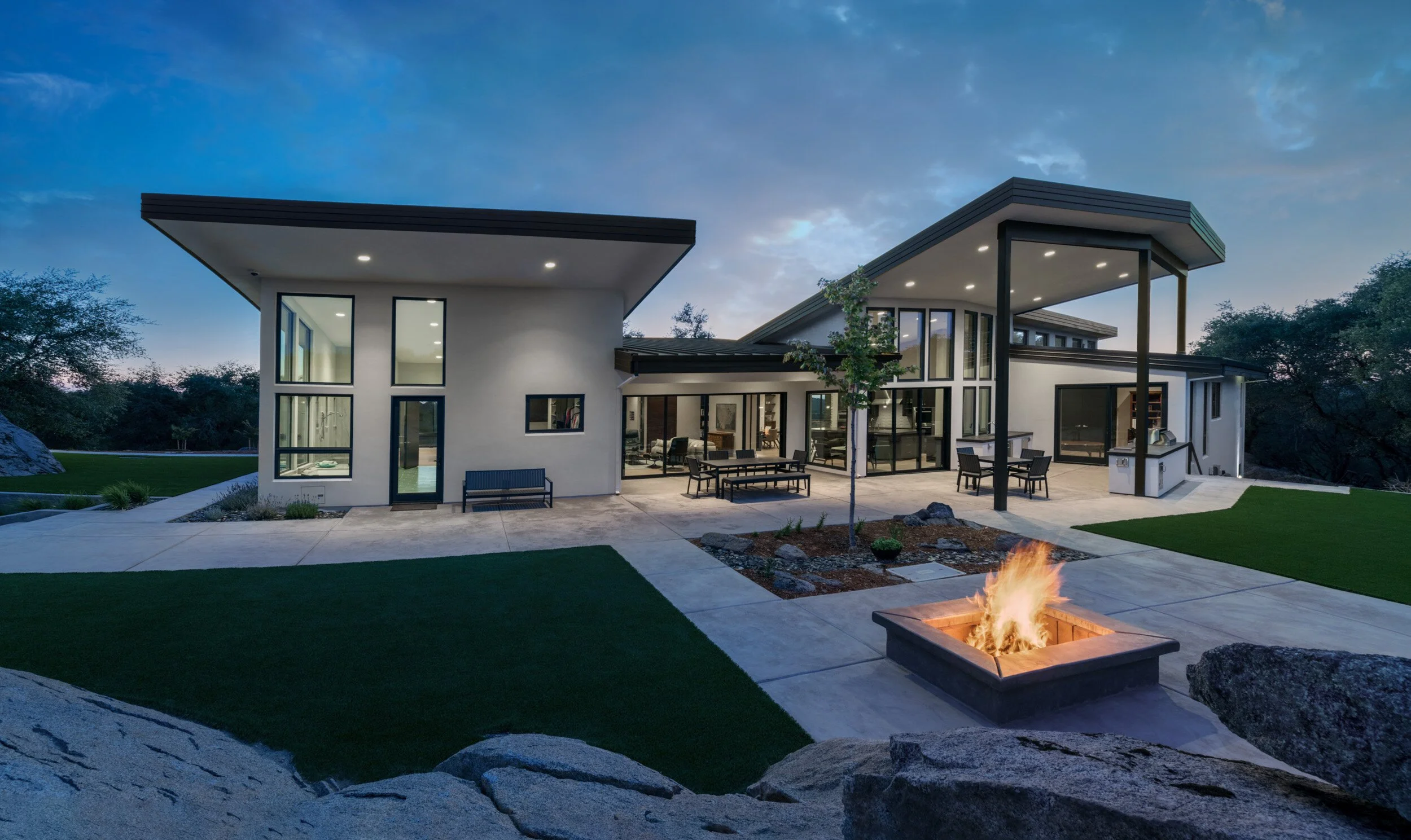 Modern house with large glass windows and a backyard patio, featuring outdoor furniture, a fire pit with a fire, and surrounded by rocks and trees at dusk.