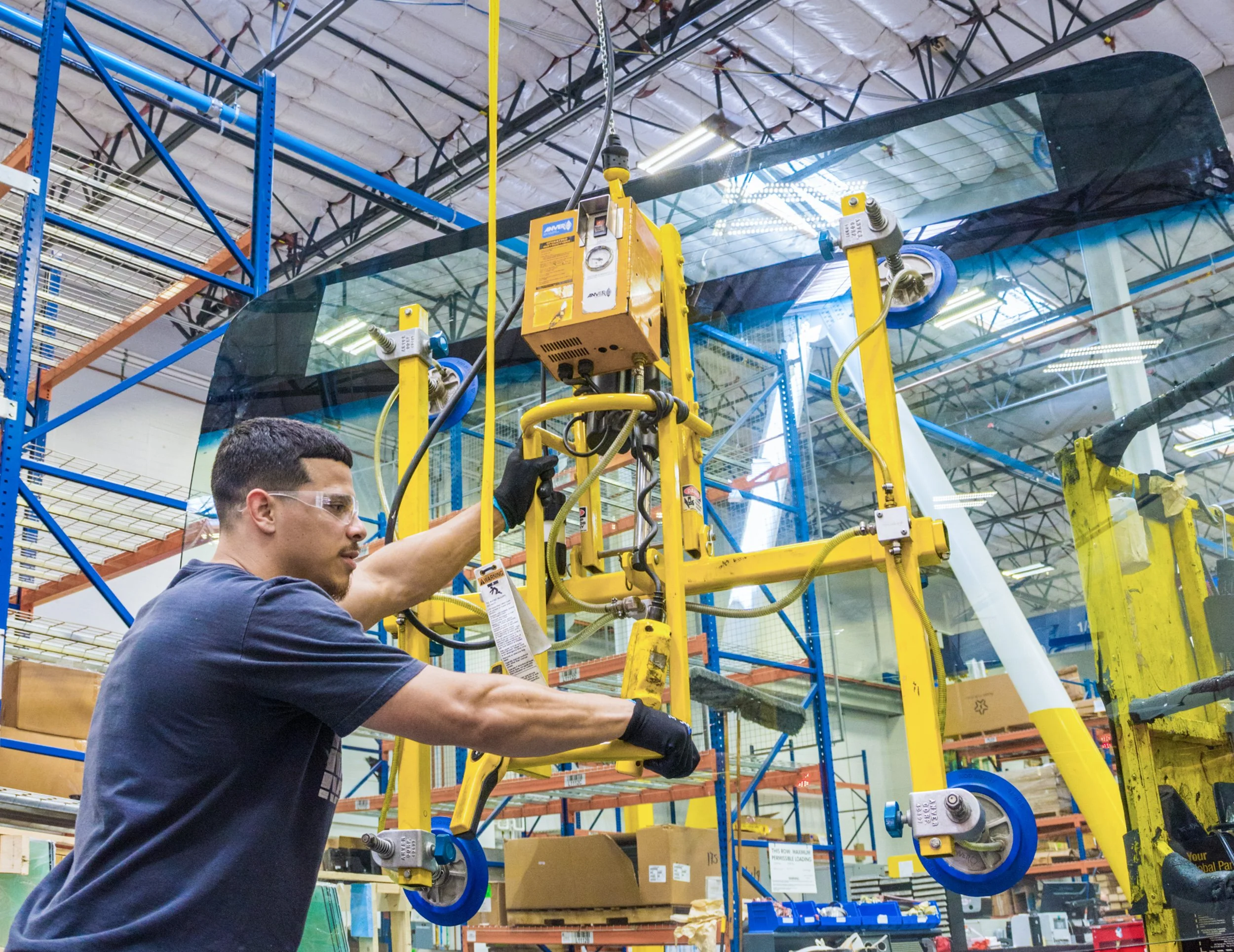 A worker in safety glasses and gloves operating machinery inside a large warehouse or manufacturing facility. The process involves handling large sheets of glass.